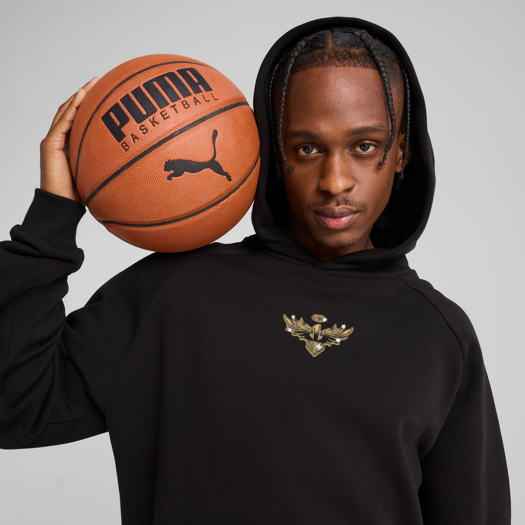 Ein Model posiert mit einem Basketball vor grauem Hintergrund, trägt ein schwarzes Kapuzenshirt.