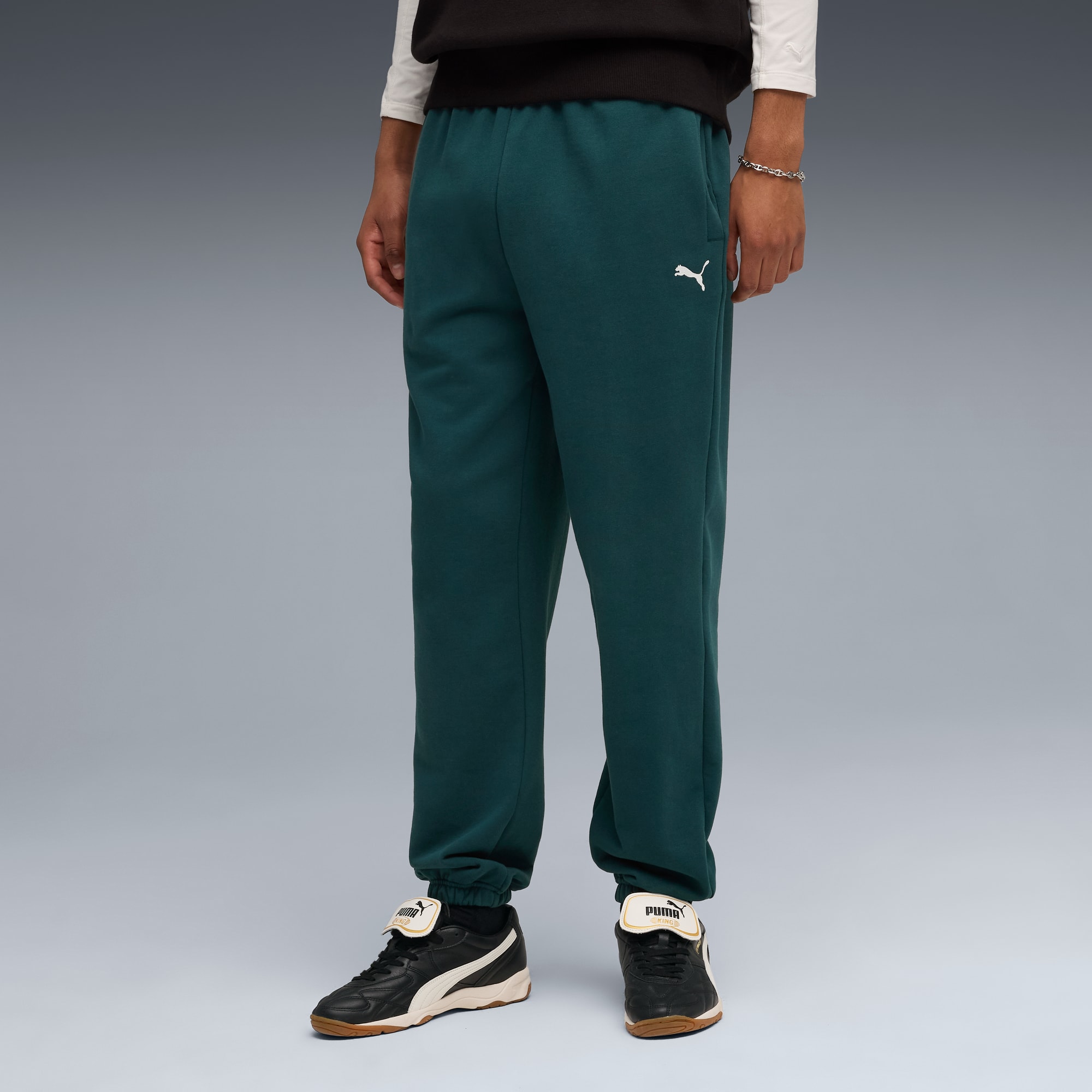 Su sfondo grigio, un modello indossa pantaloni da jogging verde scuro, scarpe da ginnastica e top nero.