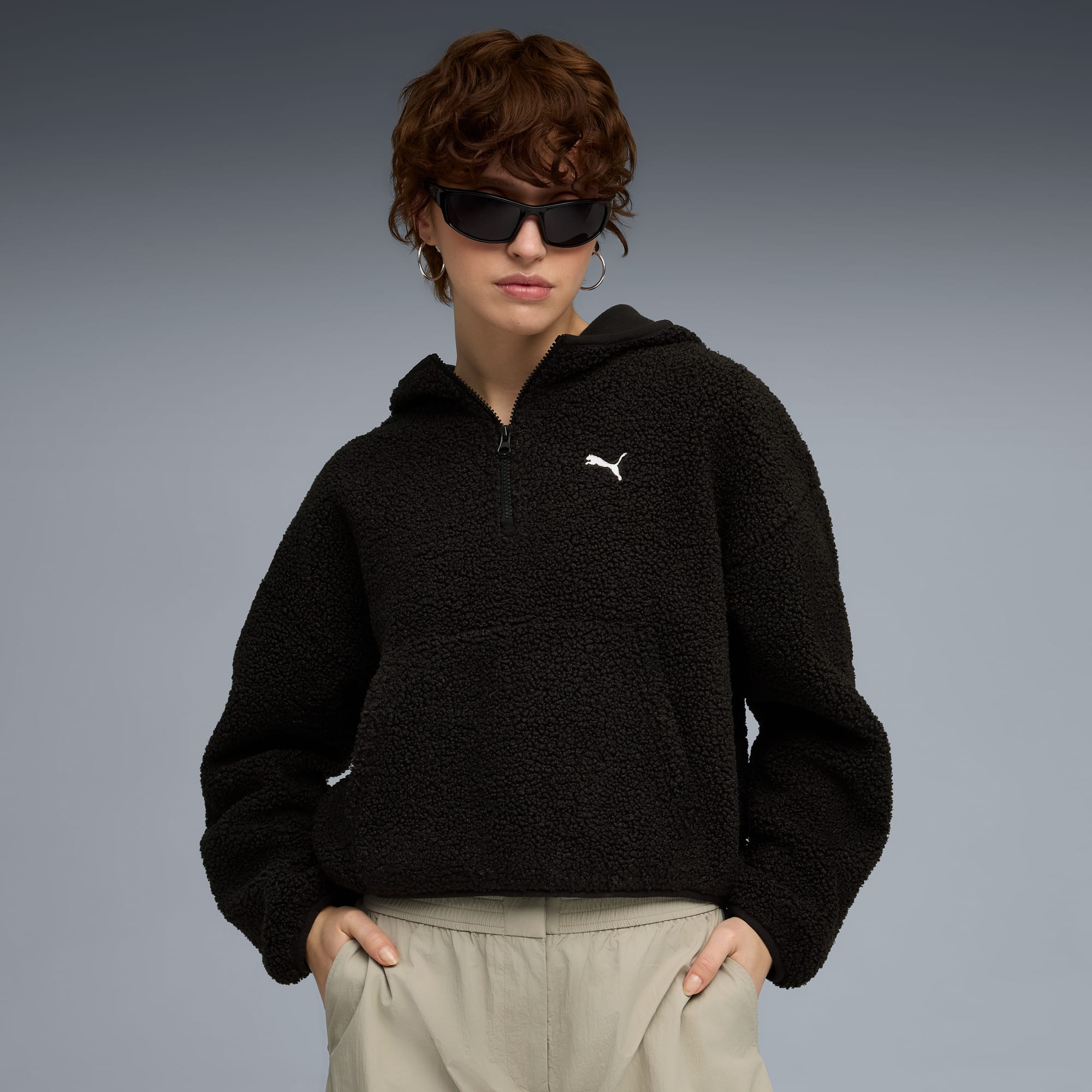 Model vor grauem Hintergrund mit dunklem, flauschigem Kapuzenpullover, Sonnenbrille und heller Hose.