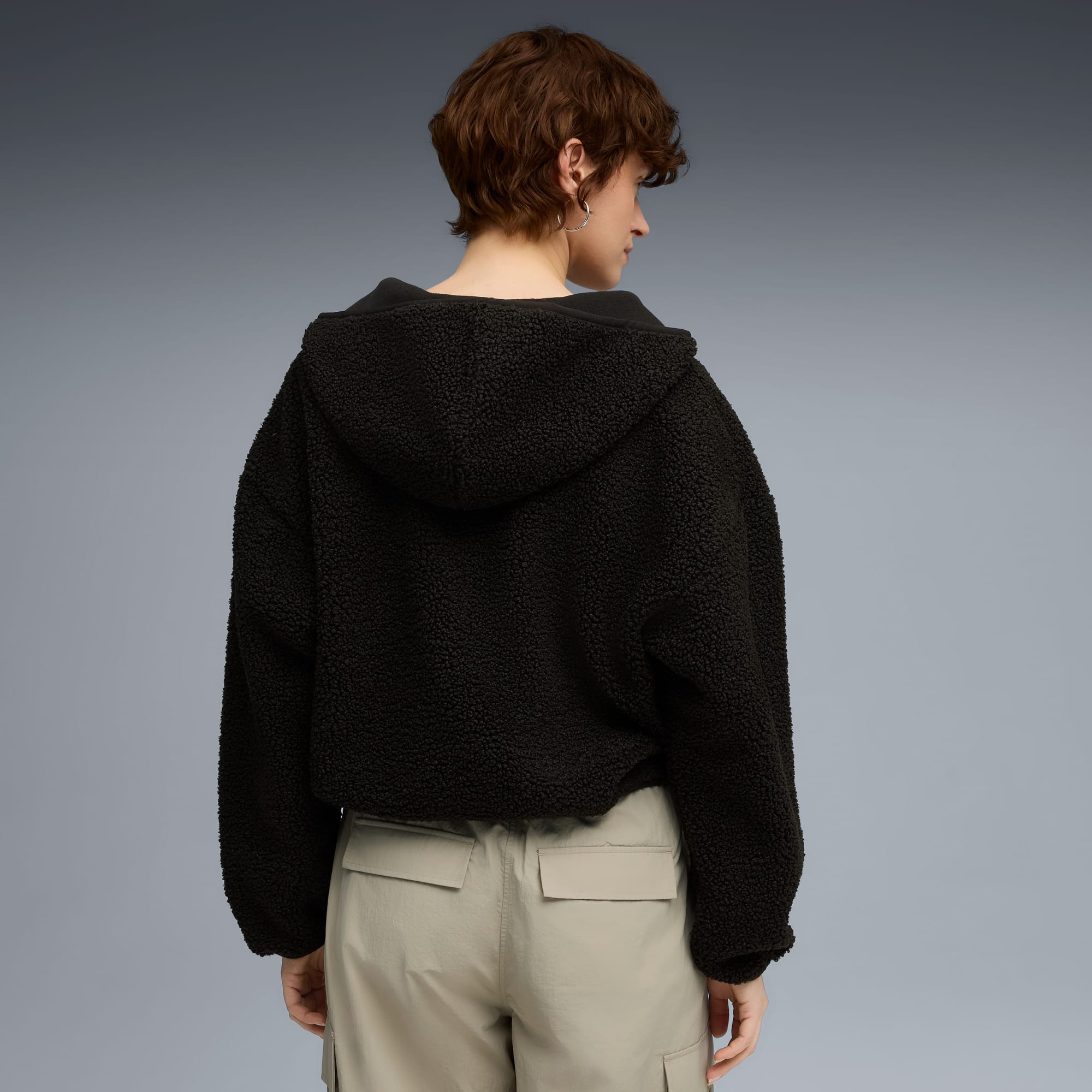 Ein Model steht vor grauem Hintergrund mit schwarzer Fleecejacke mit Kapuze und beigen Hosen.
