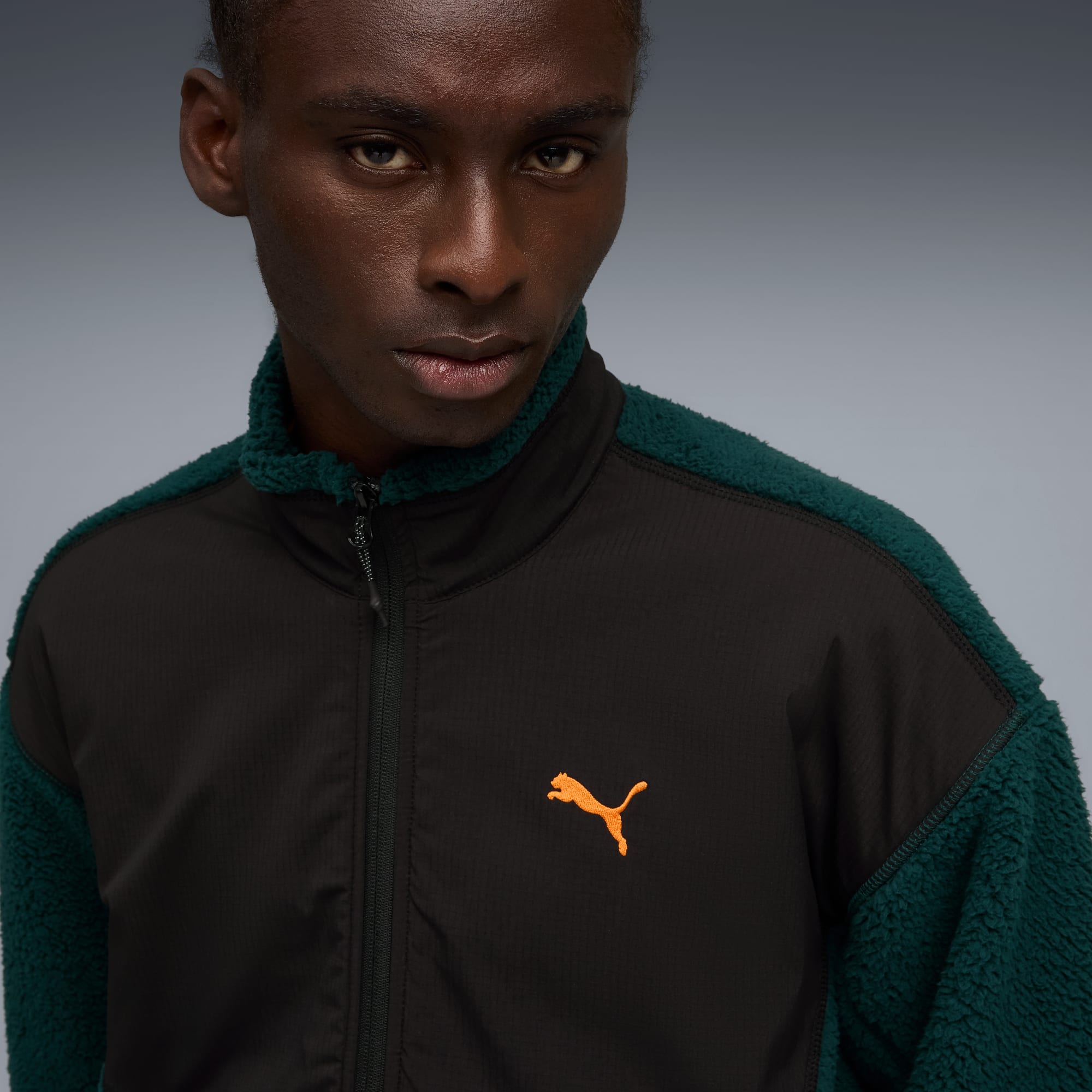 Tegen een grijze achtergrond draagt een model een jas met rits, zwart voorpand, groene fleece en oranje logo.