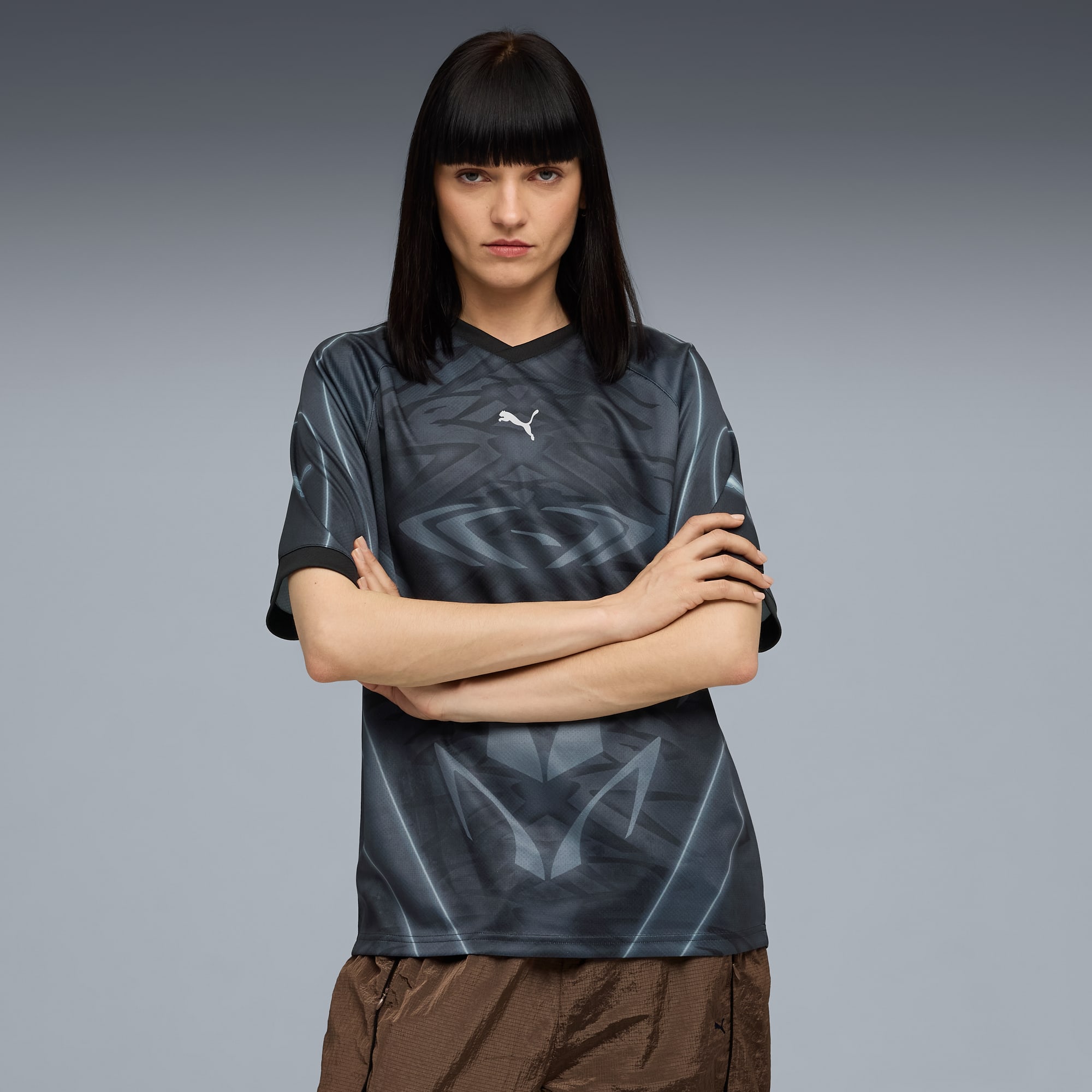 Ein Model mit verschr&auml;nkten Armen tr&auml;gt ein gemustertes Oberteil und braune Shorts vor grauem Hintergrund.