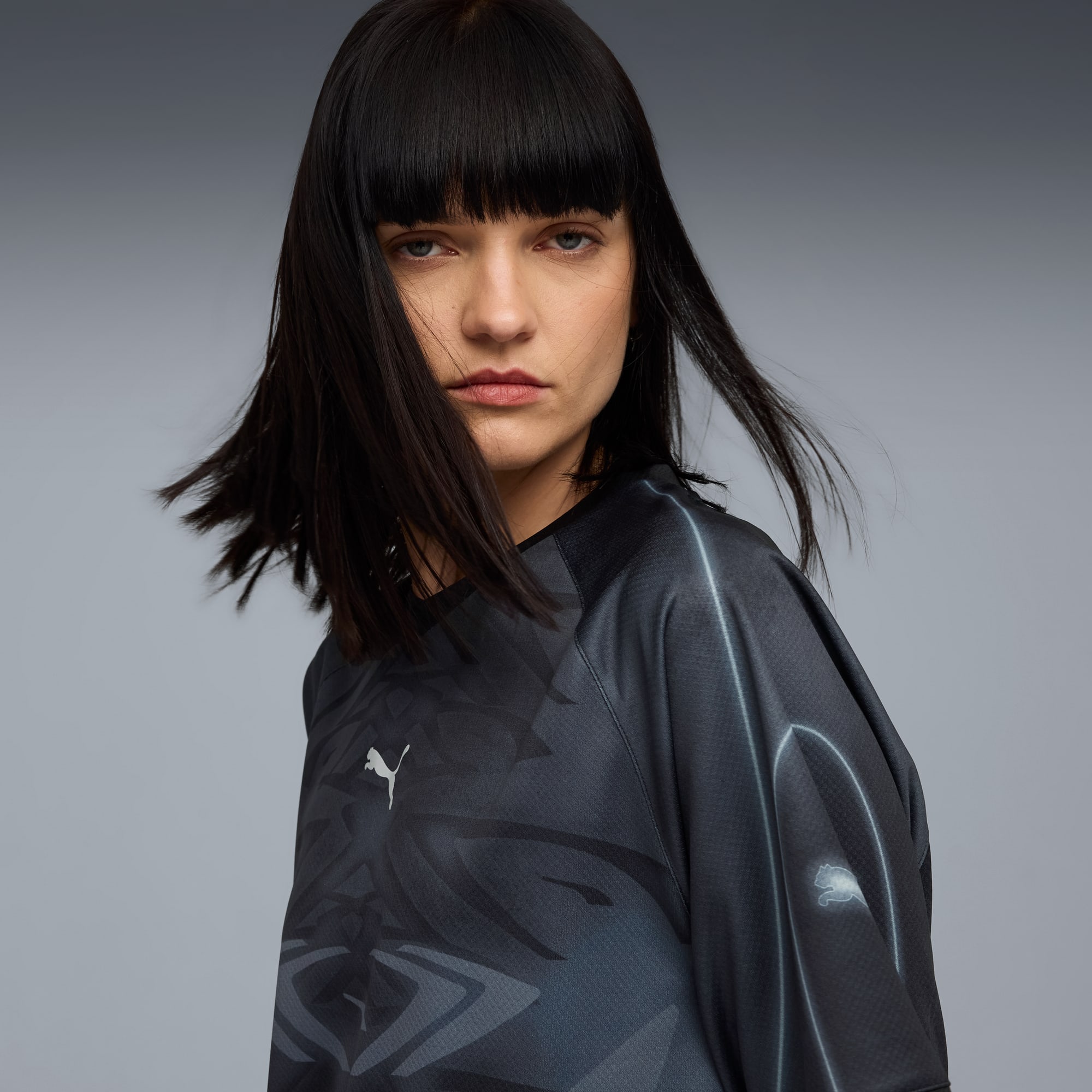 Ein Model mit dunklen, kurzen Haaren posiert vor grauem Hintergrund in einem gemusterten, dunklen Oberteil.