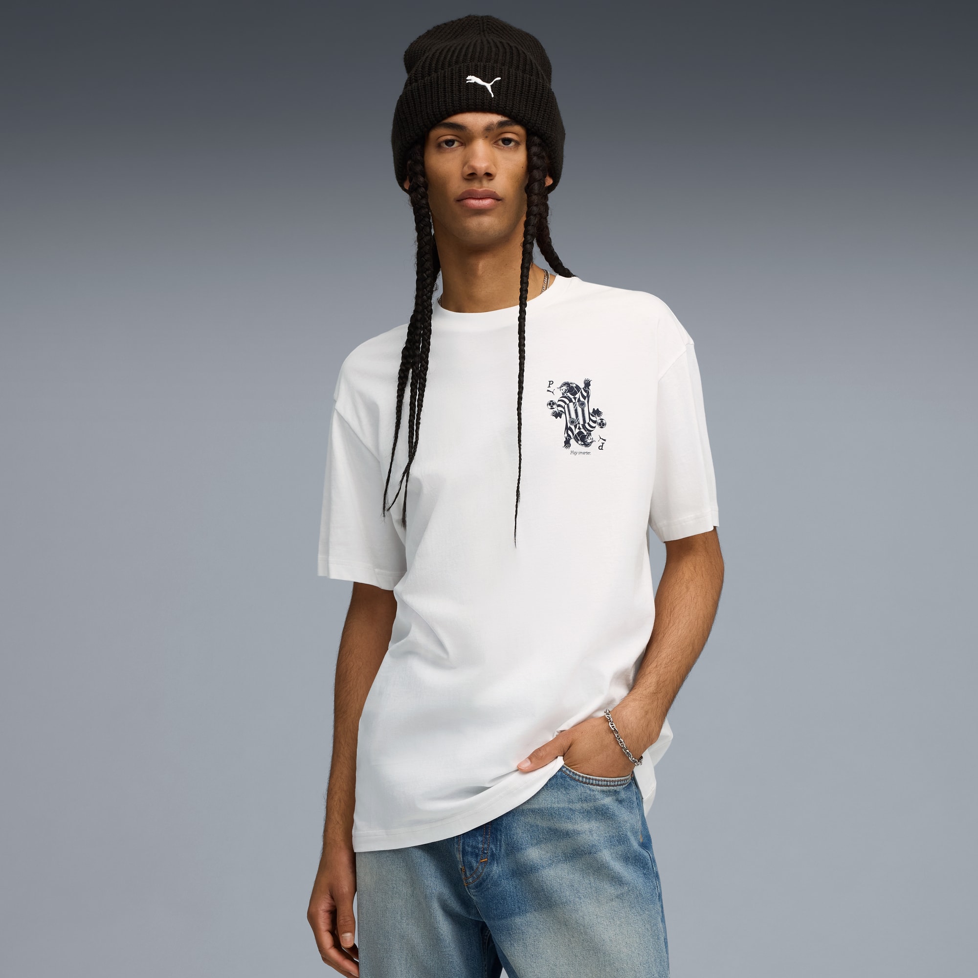 Un modello posa su sfondo grigio, indossando un berretto, una t-shirt bianca e jeans blu.