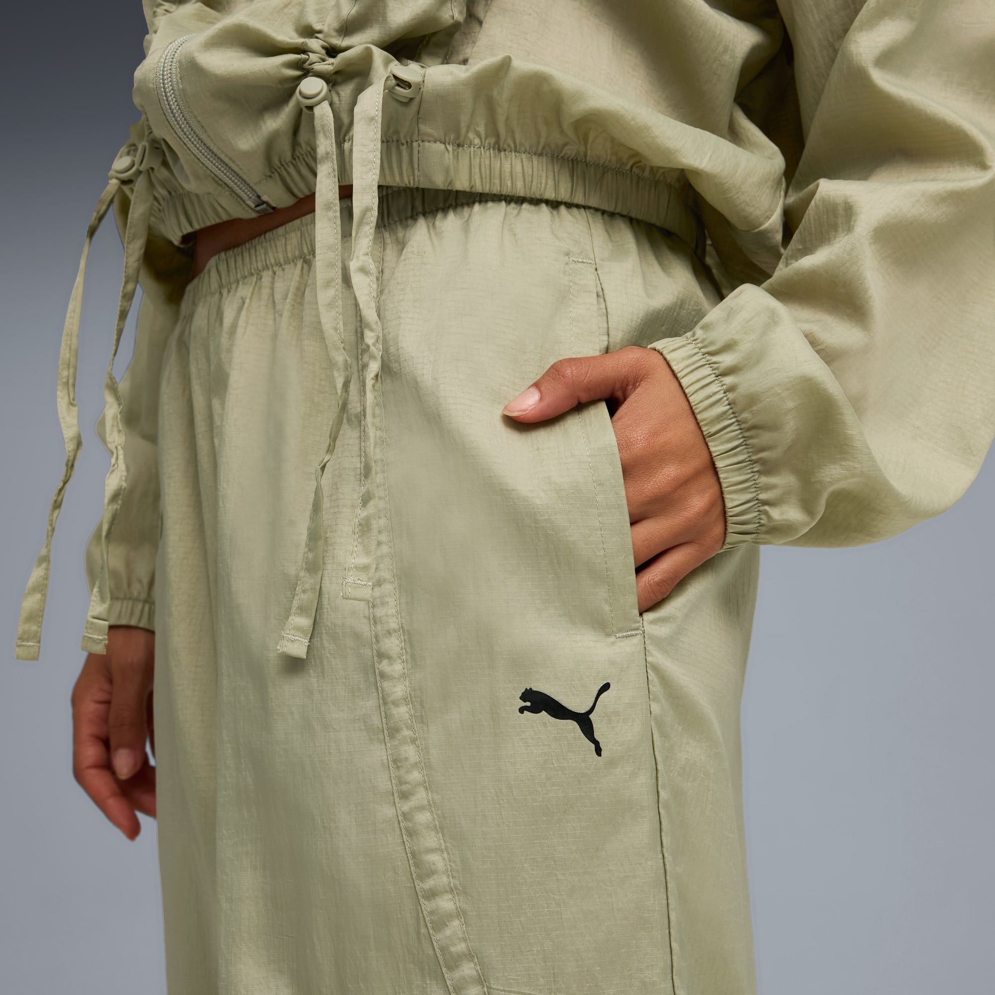 Tegen een grijze achtergrond poseert een model in een lichtgroen trainingspak met een hand in de zak.