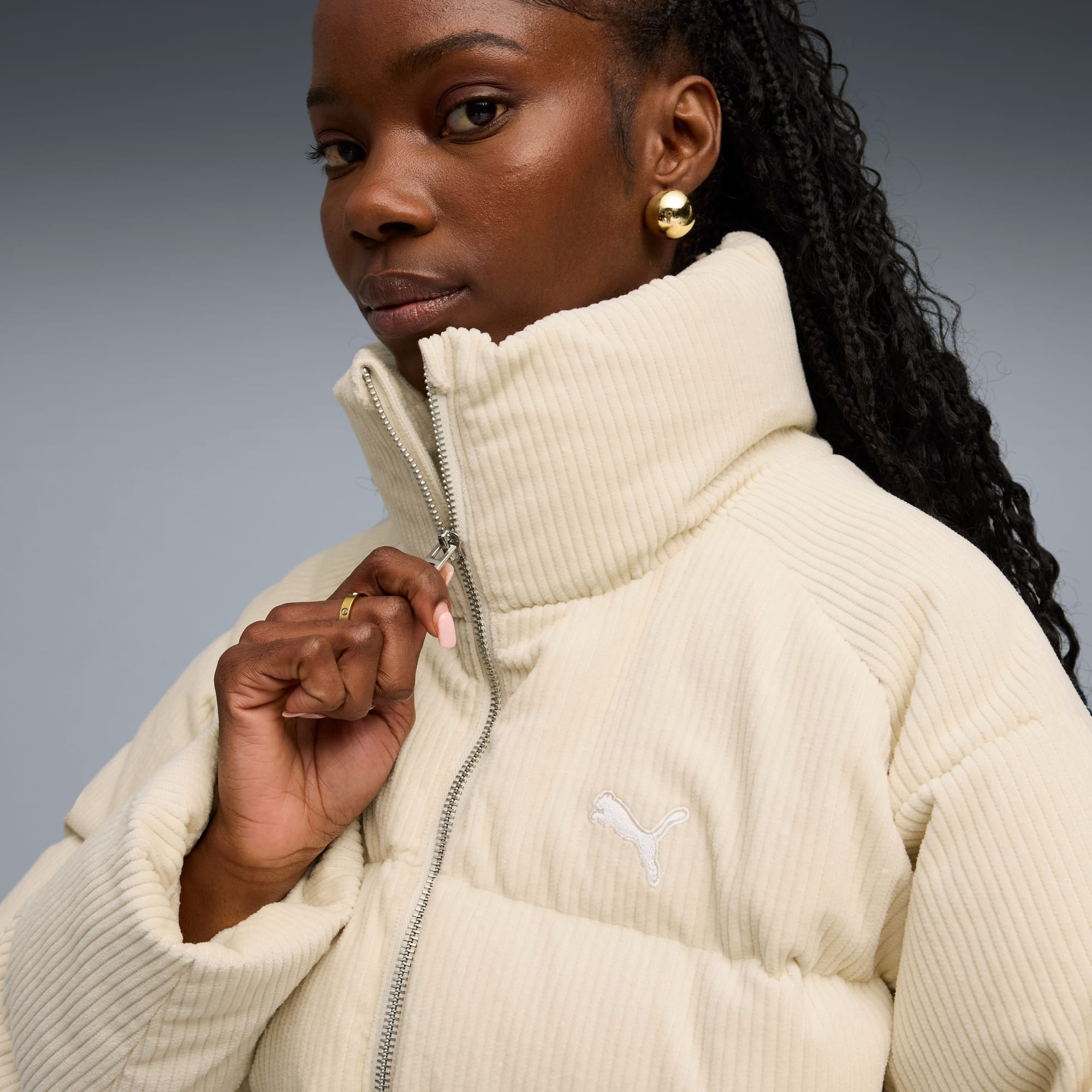 Su sfondo grigio, una modella con trecce regola la zip di un piumino in velluto a coste color crema.