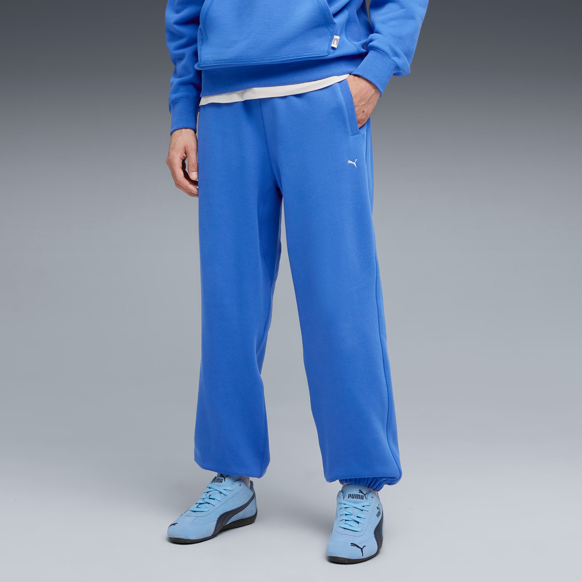 Sur fond gris, un modèle pose en pantalon de survêtement bleu et baskets assorties.