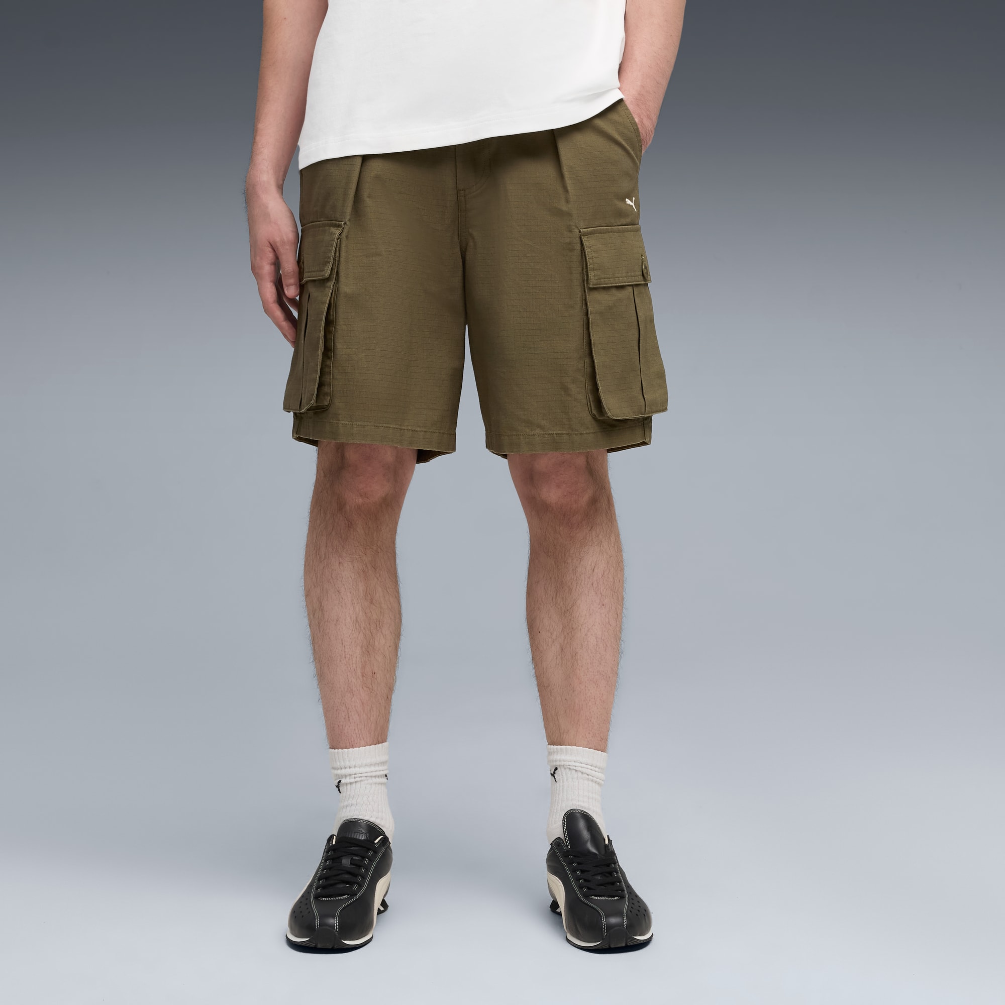 Ein Model posiert vor grau, trägt ein weißes T-Shirt, Khakishorts und schwarze Turnschuhe.