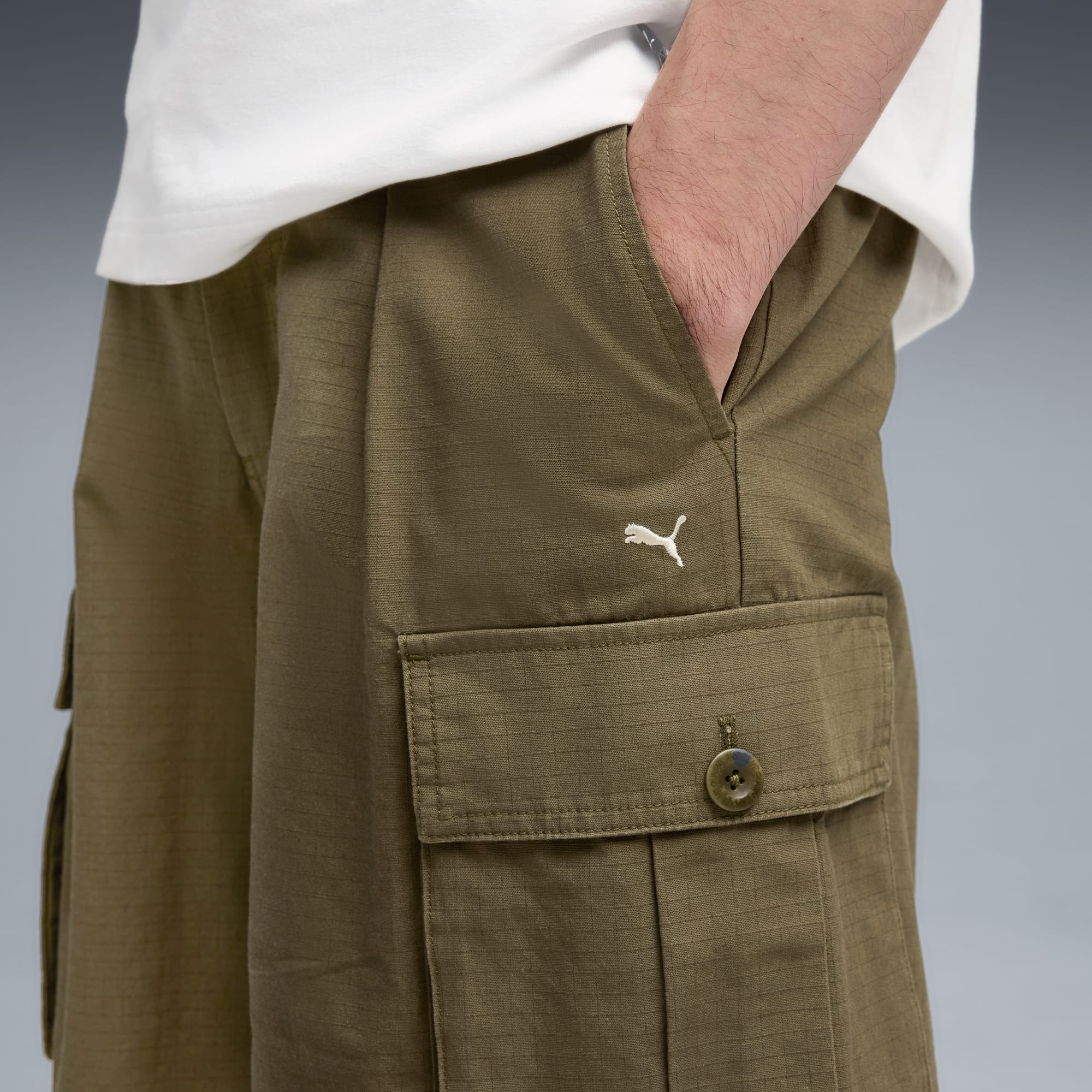 Vor grauem Hintergrund trägt eine Person olivfarbene Cargo-Shorts mit Logo und Top.