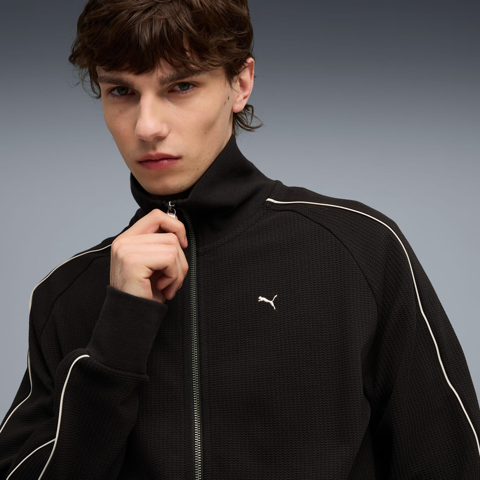 Sur fond gris, un mannequin ajuste une veste noire zippée avec passepoil et logo blancs.