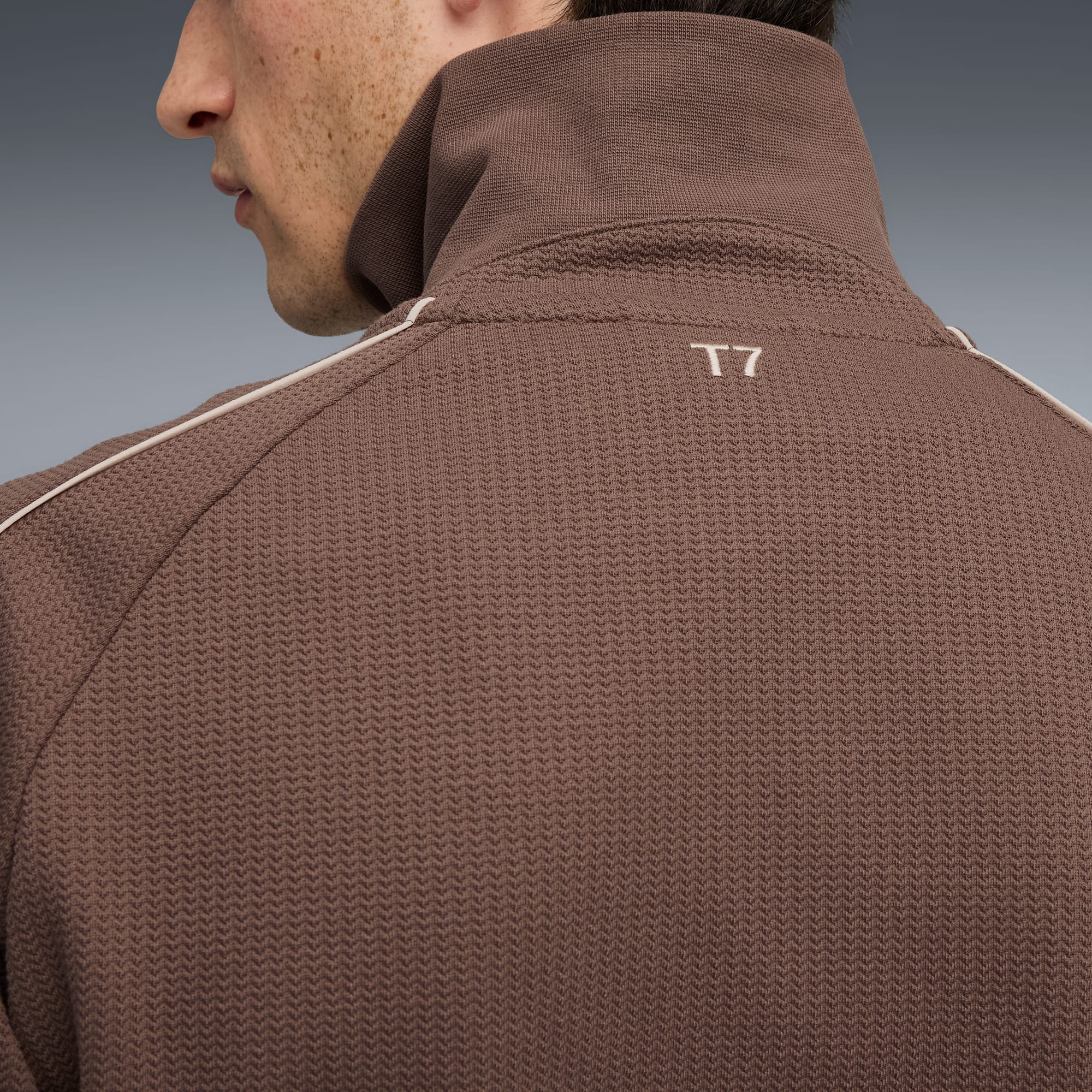 Sur un fond gris, une personne porte une veste marron texturée avec un logo 'T7'.