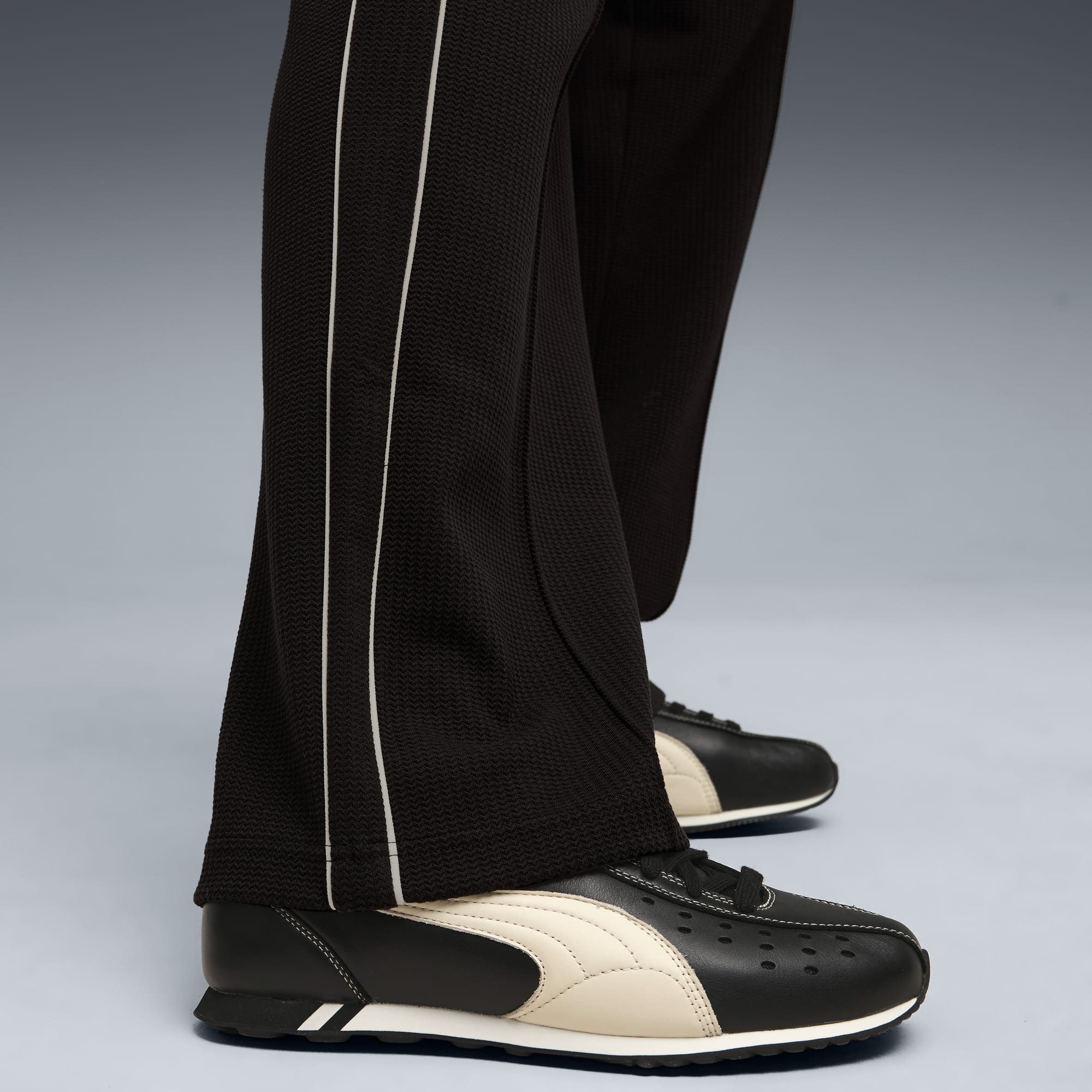Vor grauem Hintergrund trägt ein Model dunkle Hosen mit Streifen und schwarz-cremefarbene Schuhe.