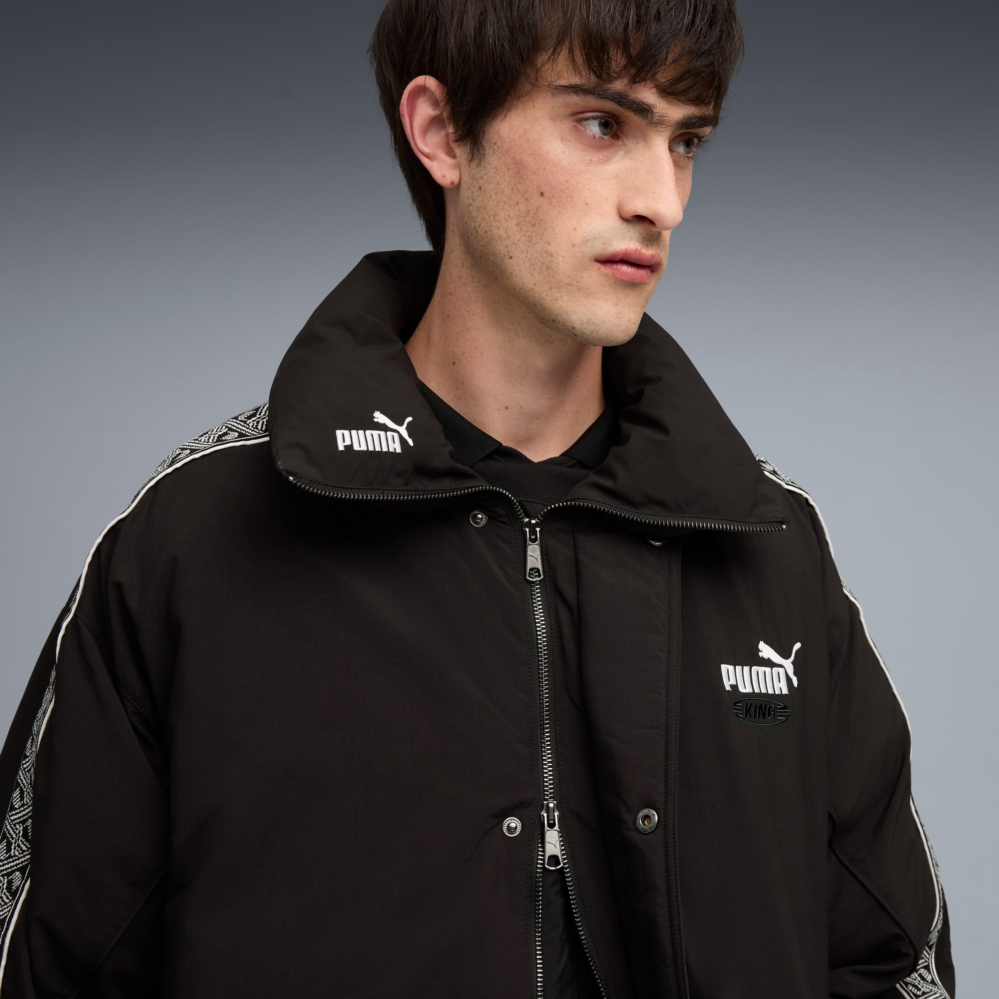 Model tegen grijze achtergrond, draagt zwarte jas met wit Puma-logo.