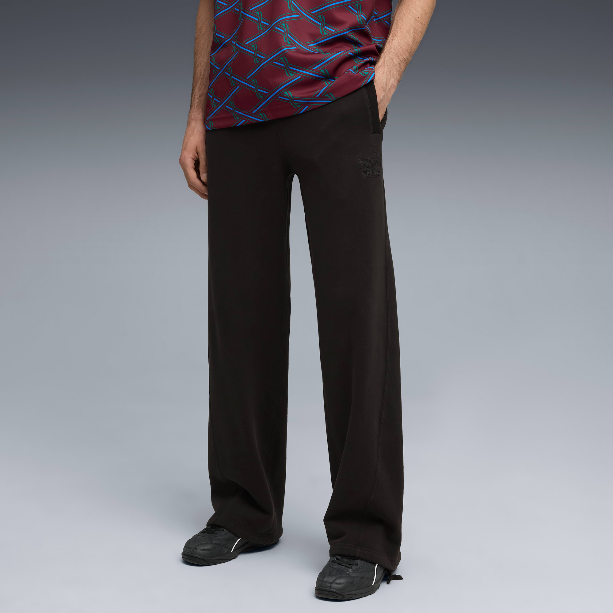 Tegen een grijze achtergrond poseert een model in een donkere broek, sportschoenen en een top met print.