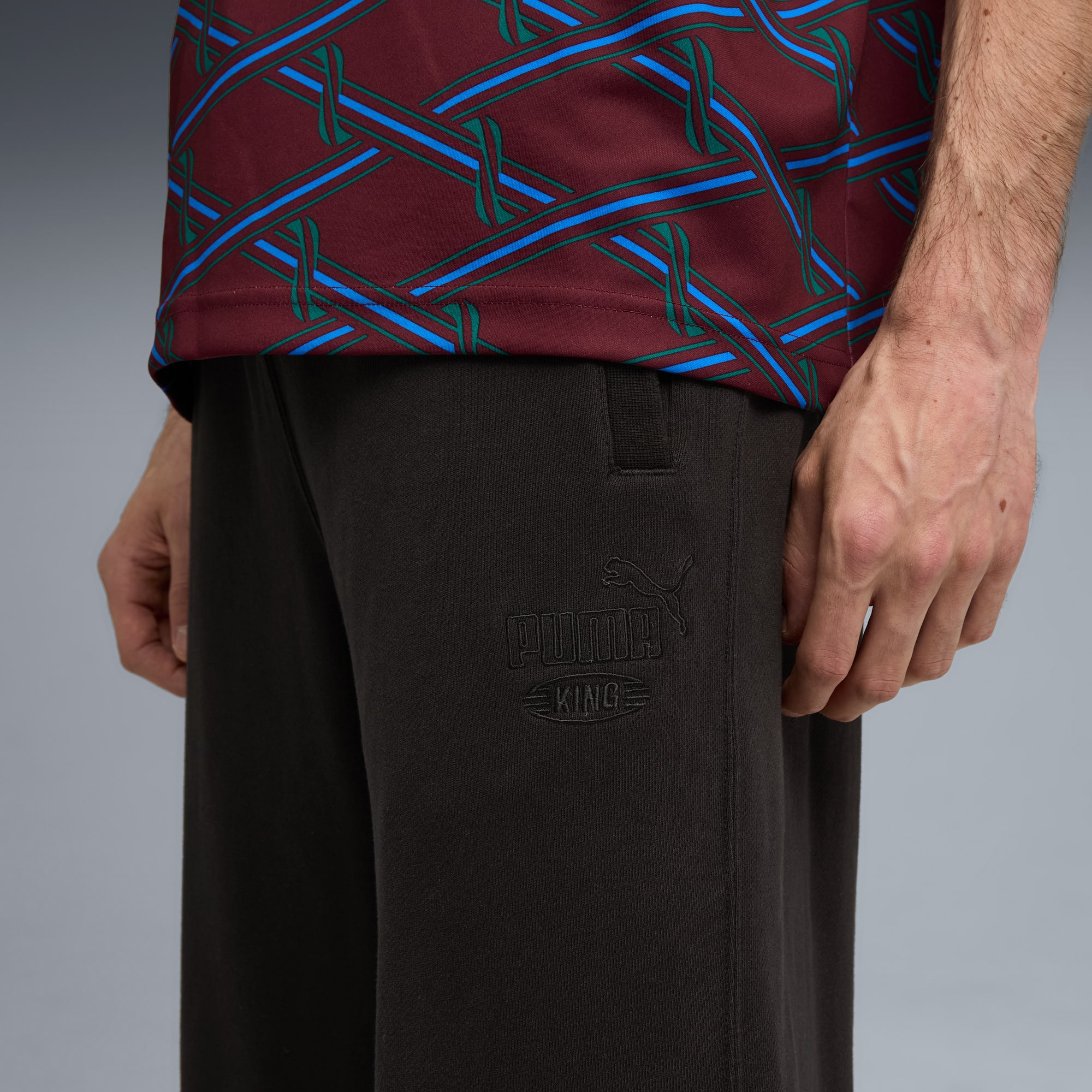 Tegen een grijze achtergrond draagt een model een top met print en een donkere 'Puma King'-broek.