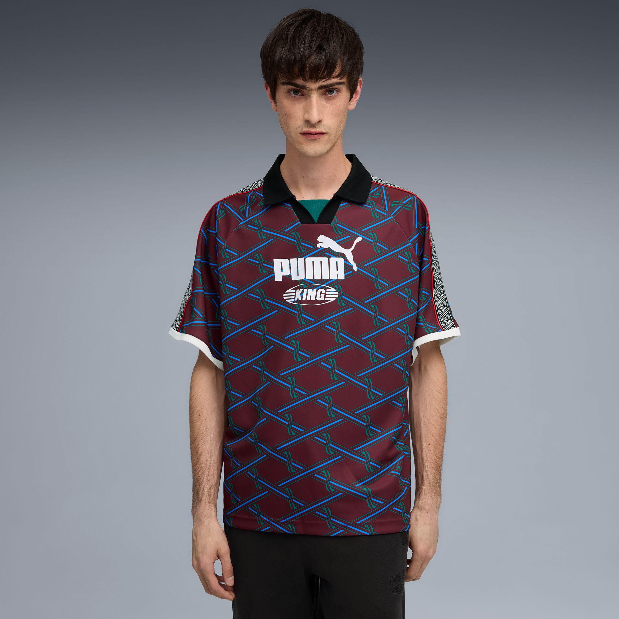 Ein Model steht vor grauem Hintergrund und trägt ein gemustertes Puma-Shirt und dunkle Hosen.