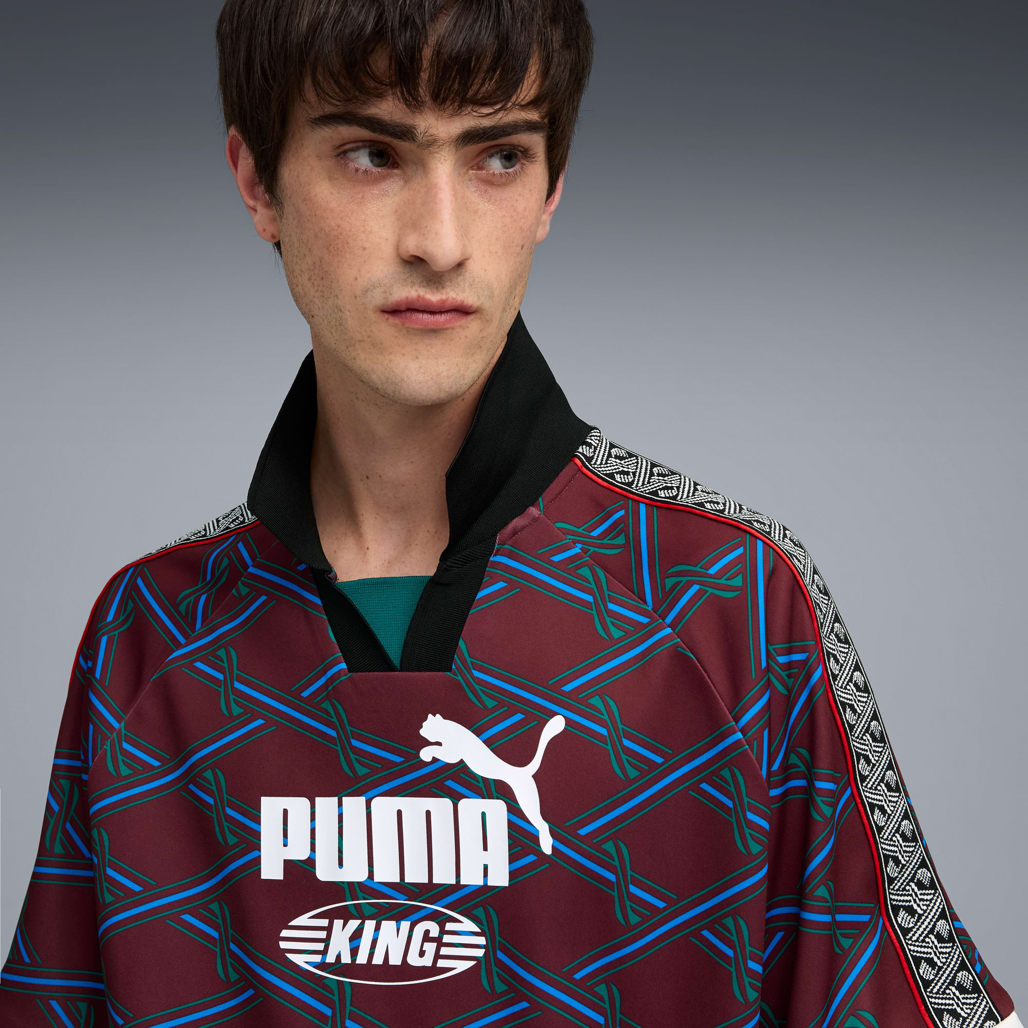 Ein Model posiert vor grauem Hintergrund mit einem gemusterten Puma King Oberteil.