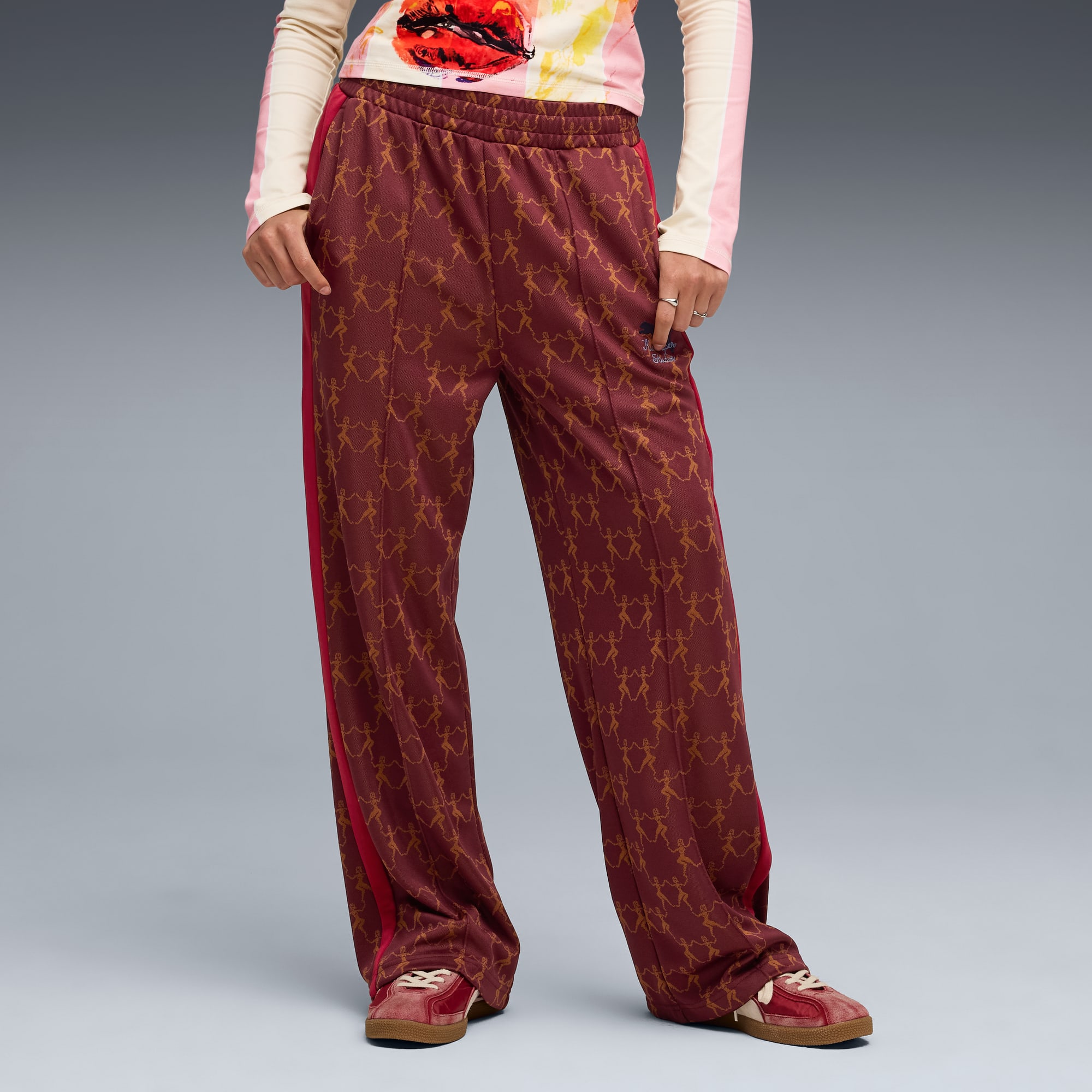 Tegen een grijze achtergrond poseert een model in een bordeauxrode broek met patroon, een top en bijpassende sneakers.