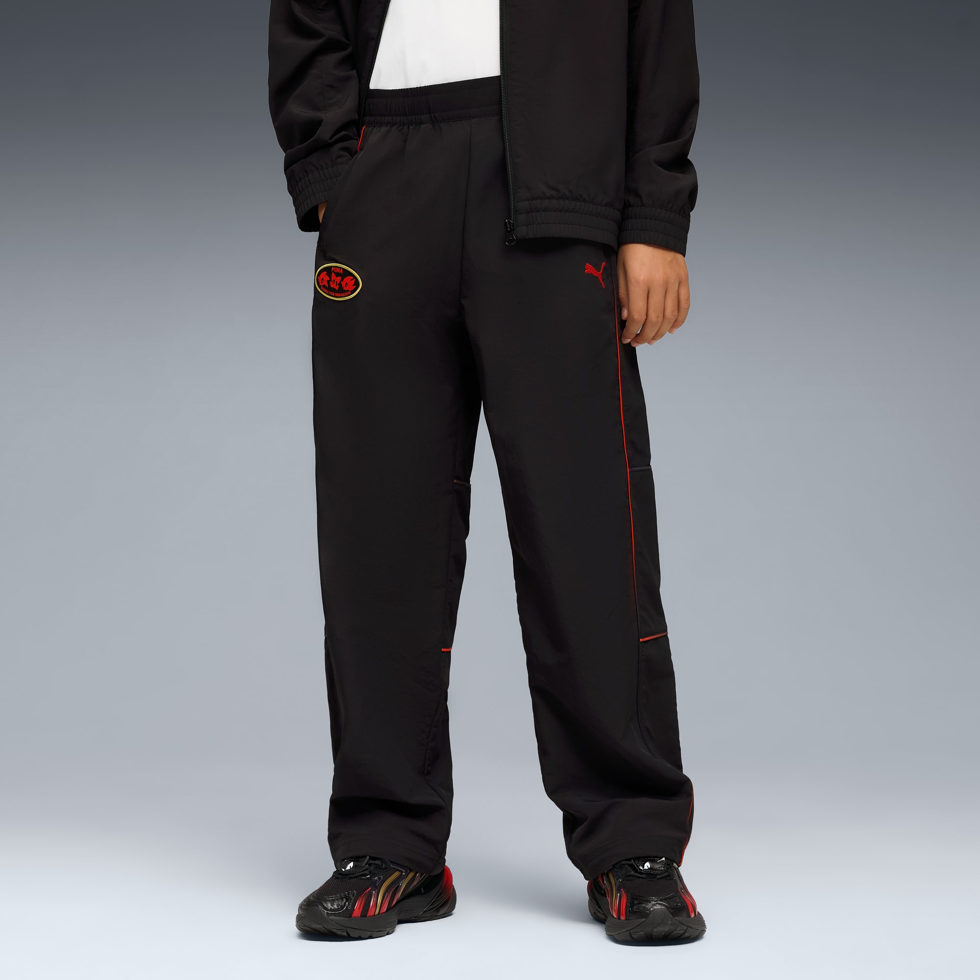 Su sfondo grigio, un modello posa indossando pantaloni neri con finiture rosse e scarpe da ginnastica abbinate.