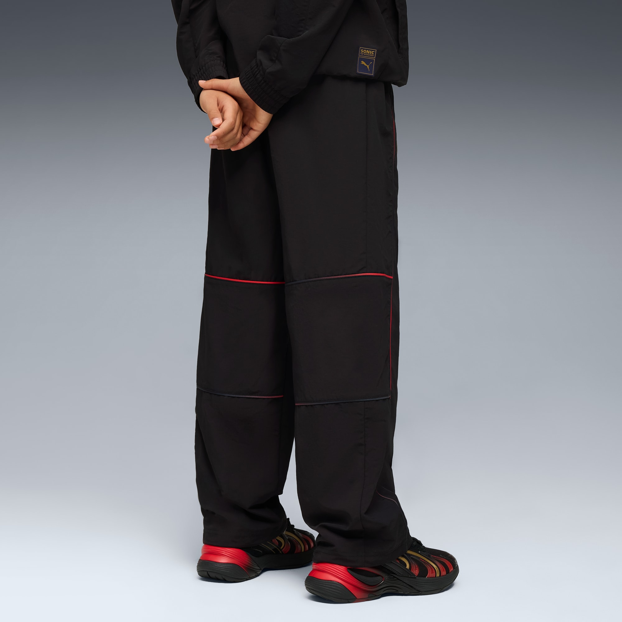 Un modello posa su sfondo grigio, indossa pantaloni neri, giacca e scarpe da ginnastica rosse e nere.