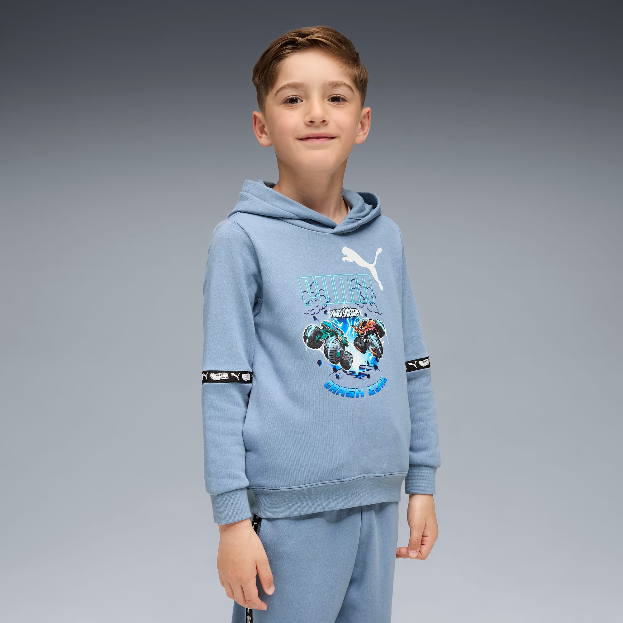 Ein Kind posiert vor grauem Hintergrund, trägt einen blauen Kapuzenpullover und Jogginghose.
