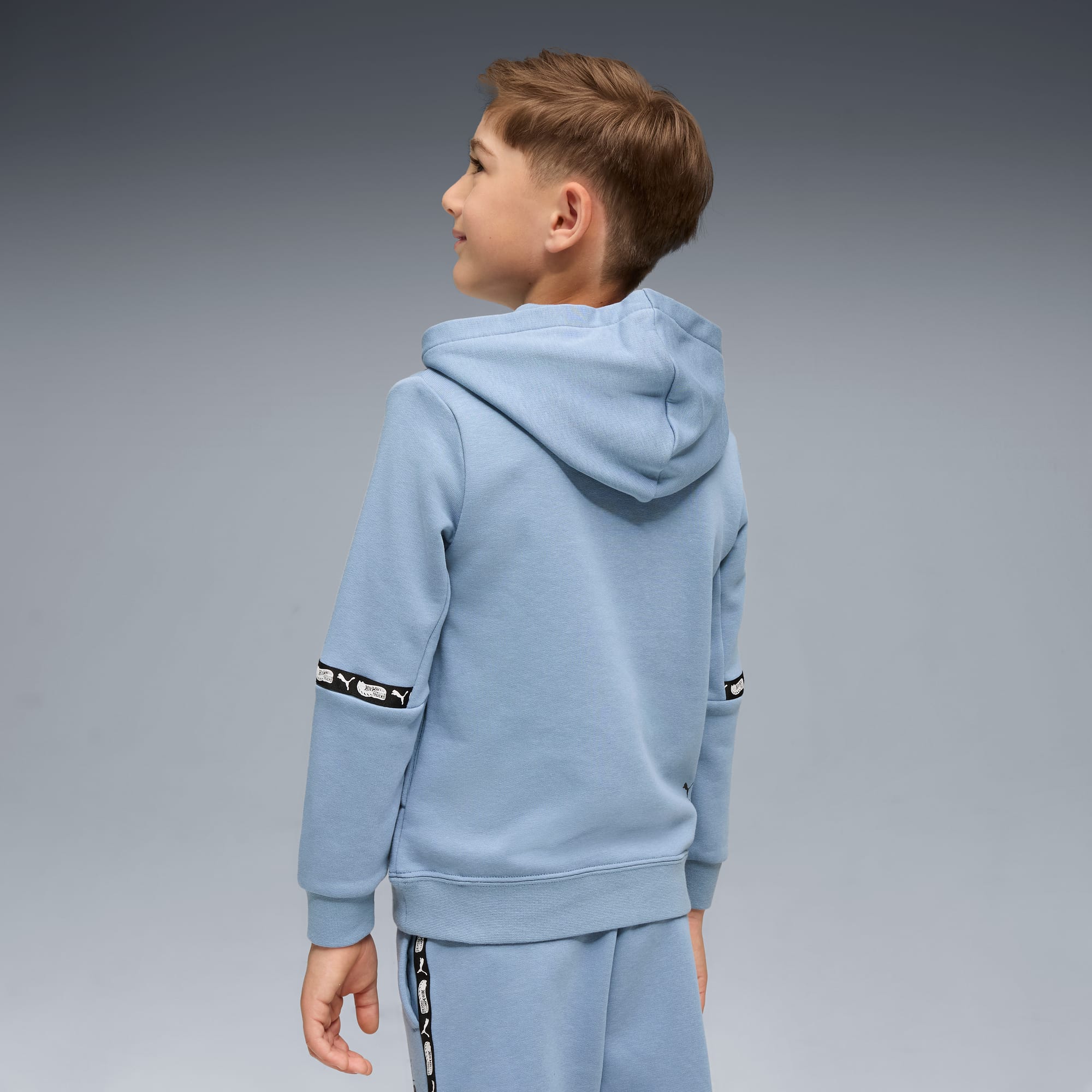 Ein Kind blickt vor grauem Hintergrund nach links, trägt einen blauen Kapuzenpullover und passende Hose.