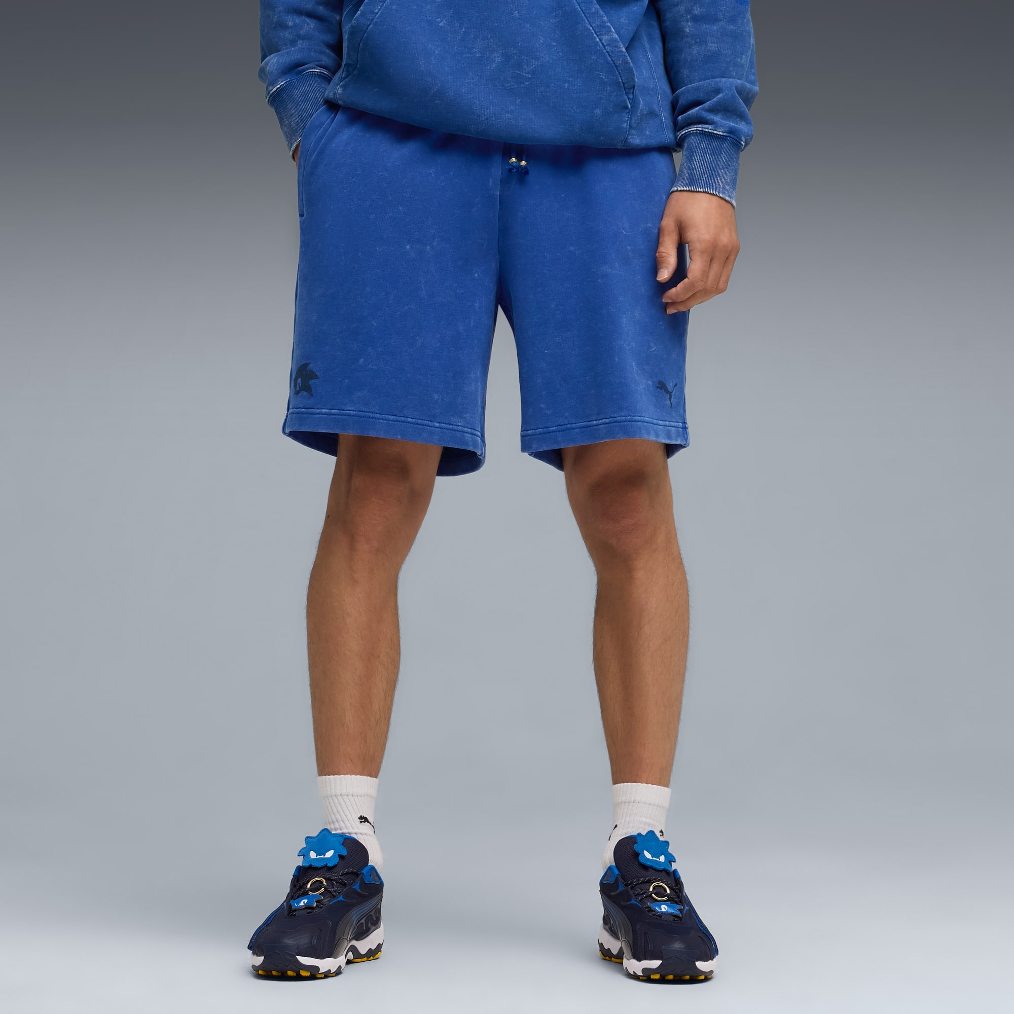 Against grey, a model wears blue shorts, a hoodie, white socks and patterned trainers.