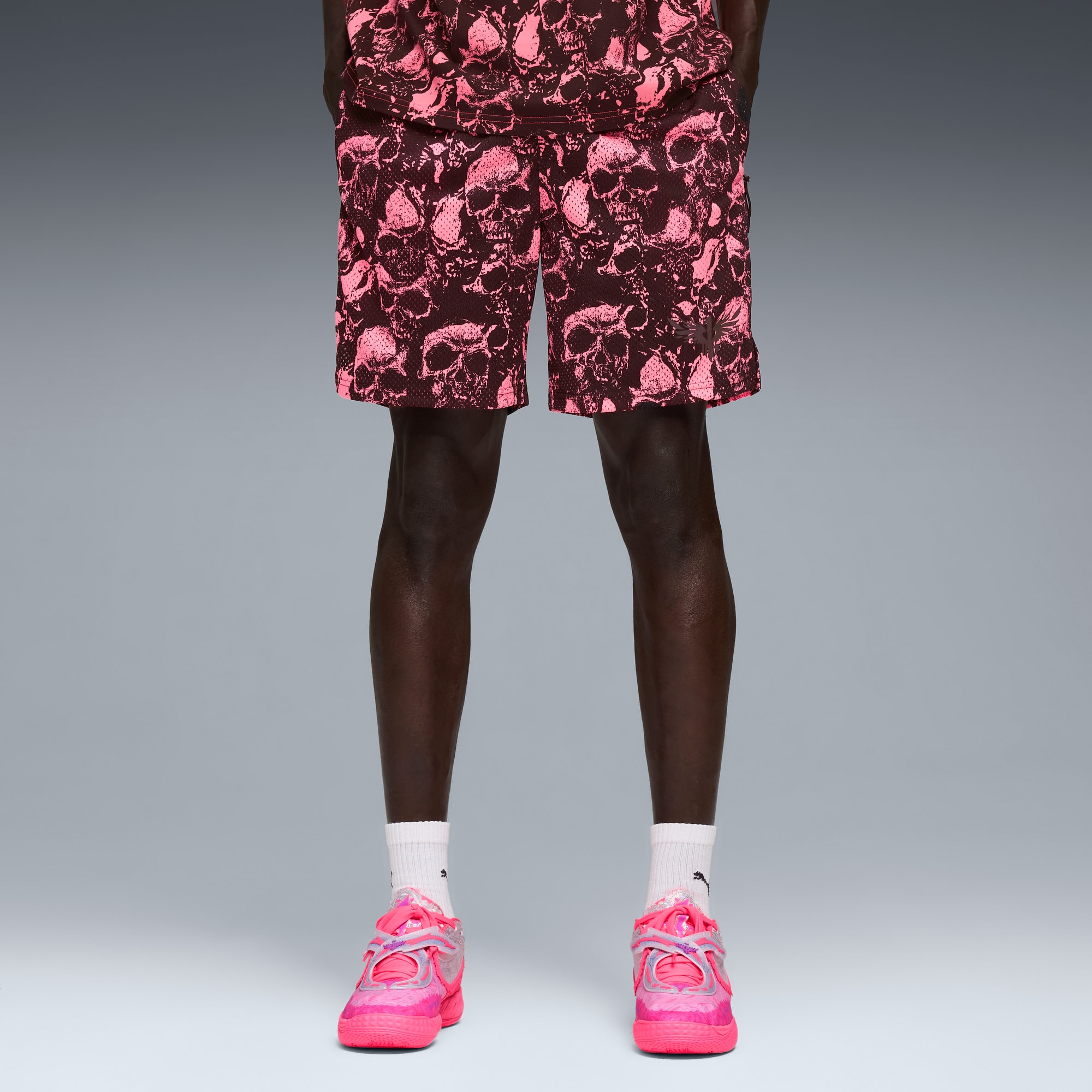 Su sfondo grigio, un modello indossa pantaloncini con stampa di teschi e scarpe da ginnastica rosa acceso con calzini bianchi.