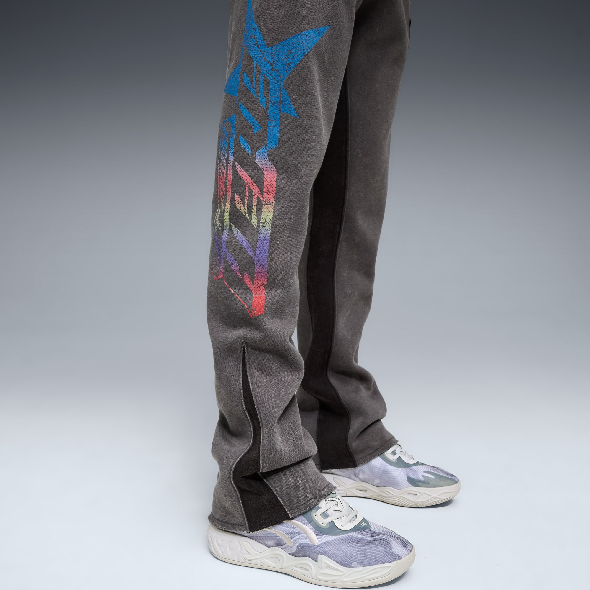 Su sfondo grigio, un modello indossa pantaloni da jogging grigi con grafiche colorate e scarpe da ginnastica fantasia.