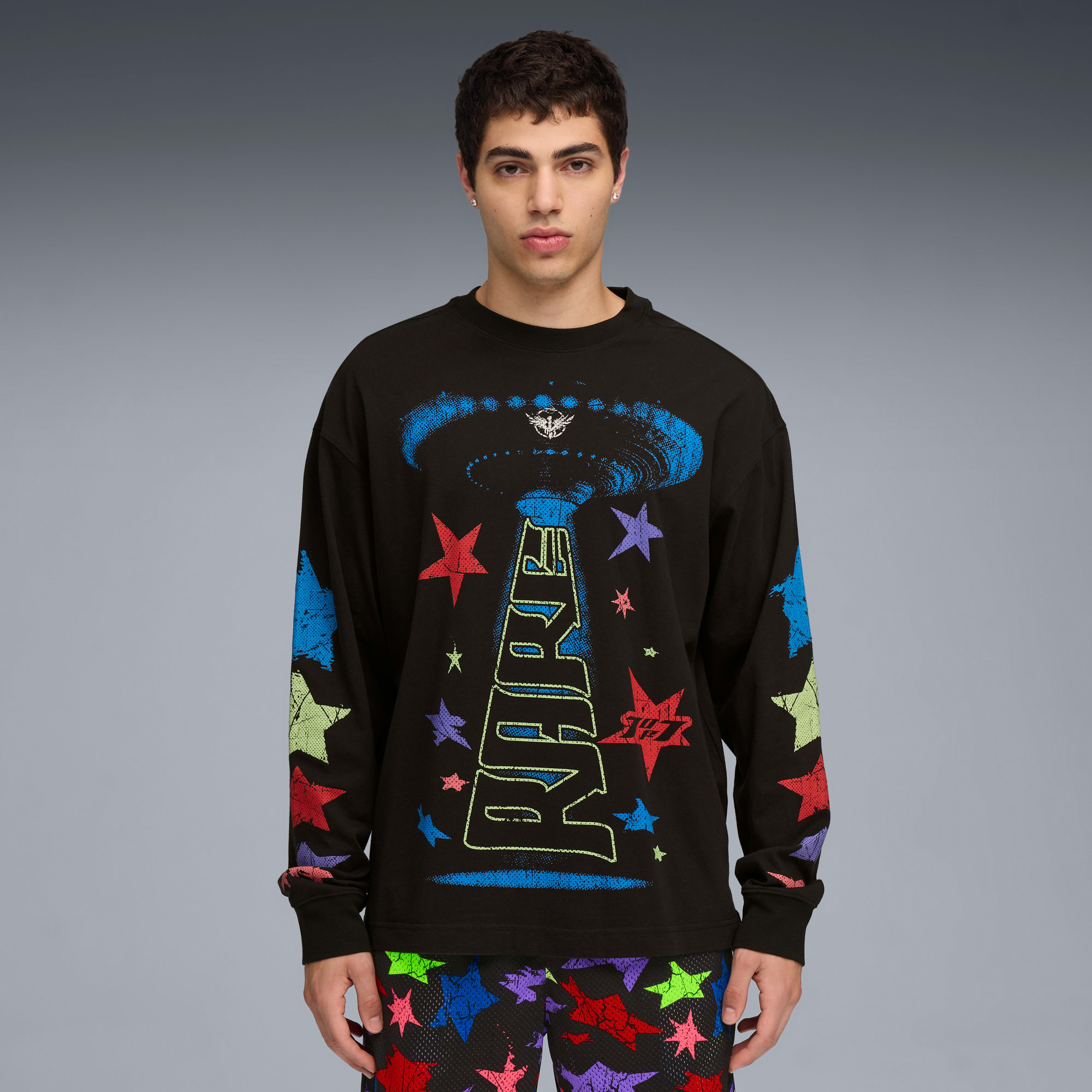 Model pozuje na szarym tle, ubrany w czarną koszulkę z długim rękawem i wzorzyste szorty.