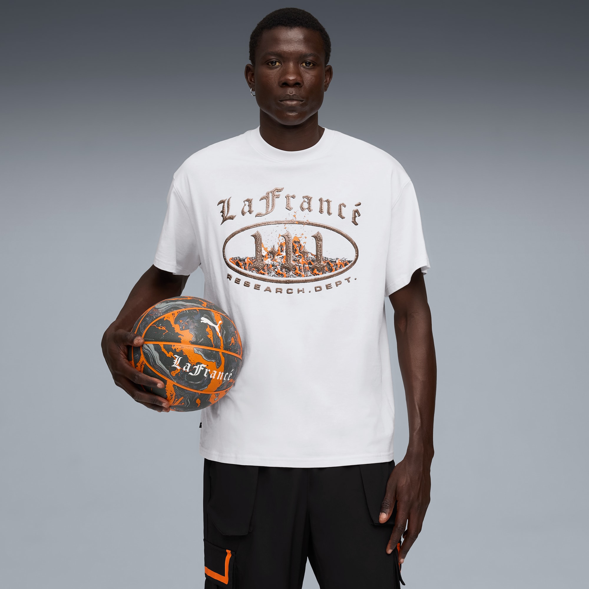 Un mannequin tient un ballon de basket, porte un t-shirt graphique et un pantalon noir sur fond gris.