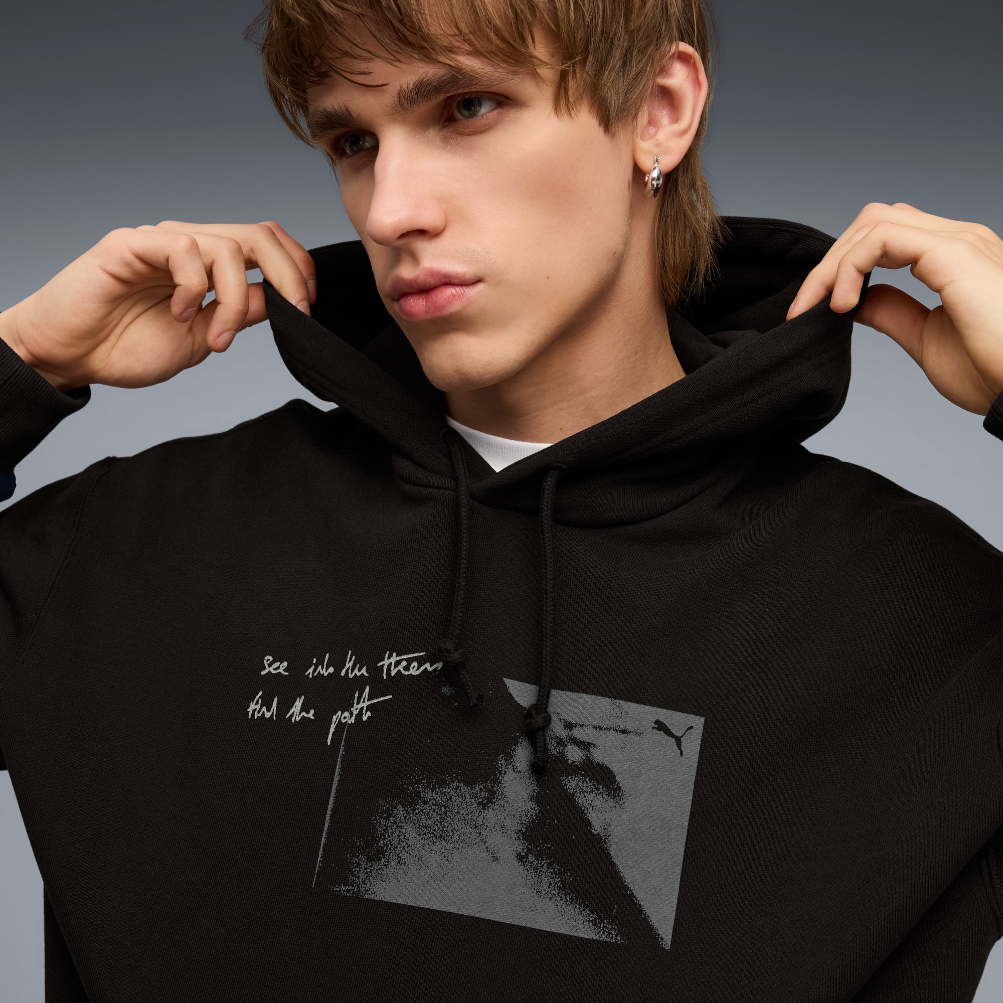 Een model past een zwarte hoodie met print aan tegen een grijze achtergrond.