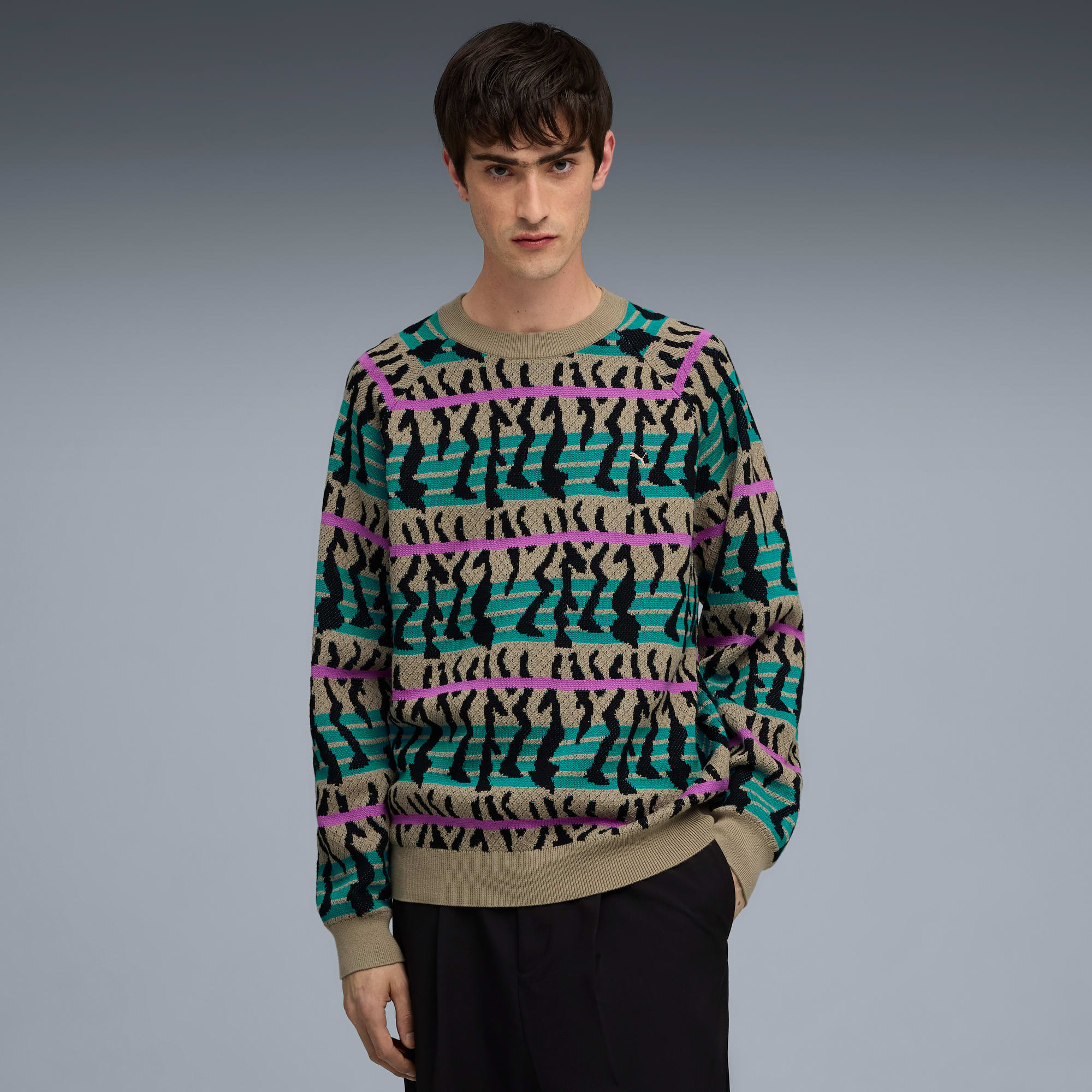 Un modello posa su sfondo grigio, indossa un maglione fantasia e pantaloni scuri.
