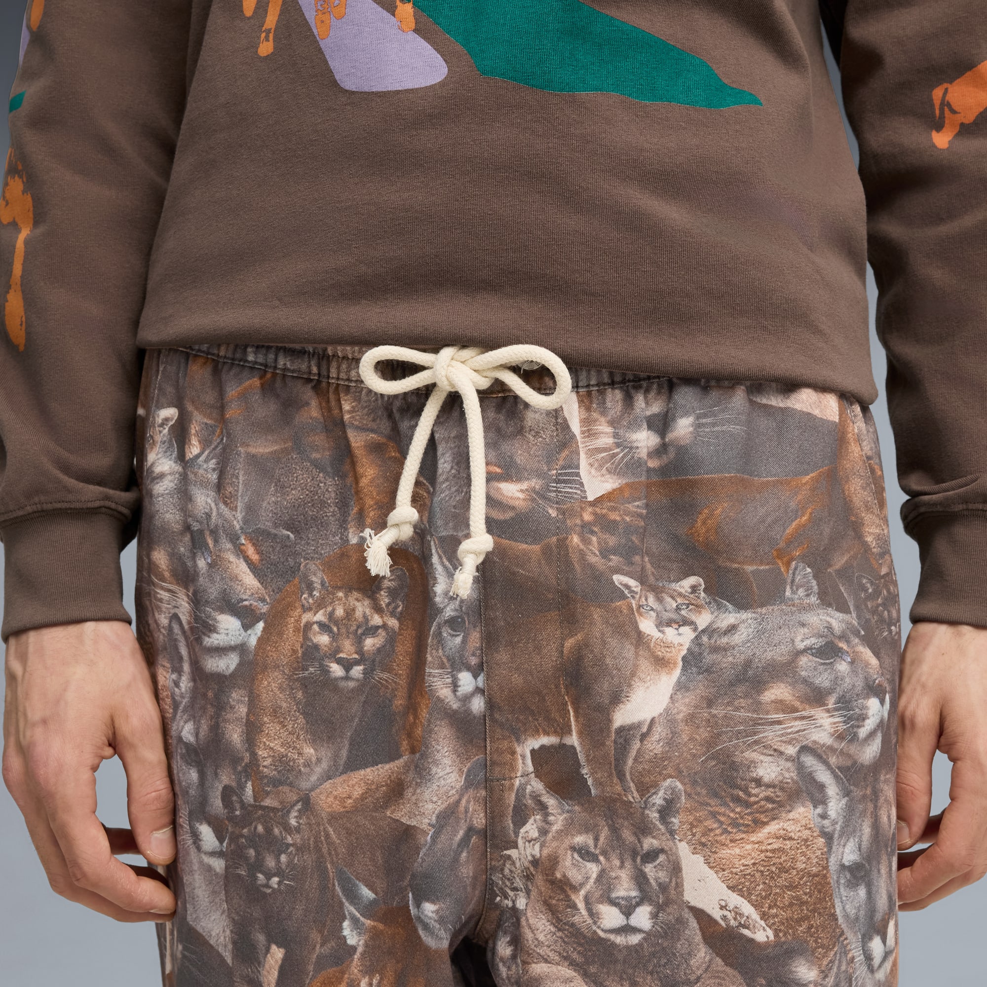 Une personne portant un haut marron et un pantalon à motif de couguars sur fond gris.