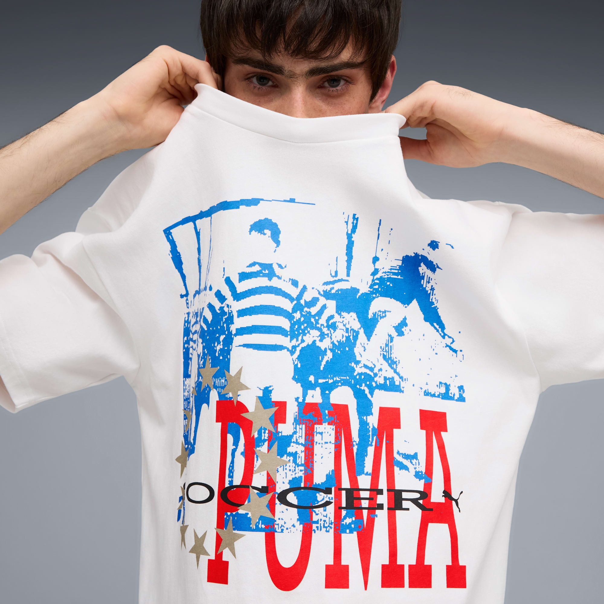 Su sfondo grigio, modello tiene una t-shirt bianca grafica davanti al viso.