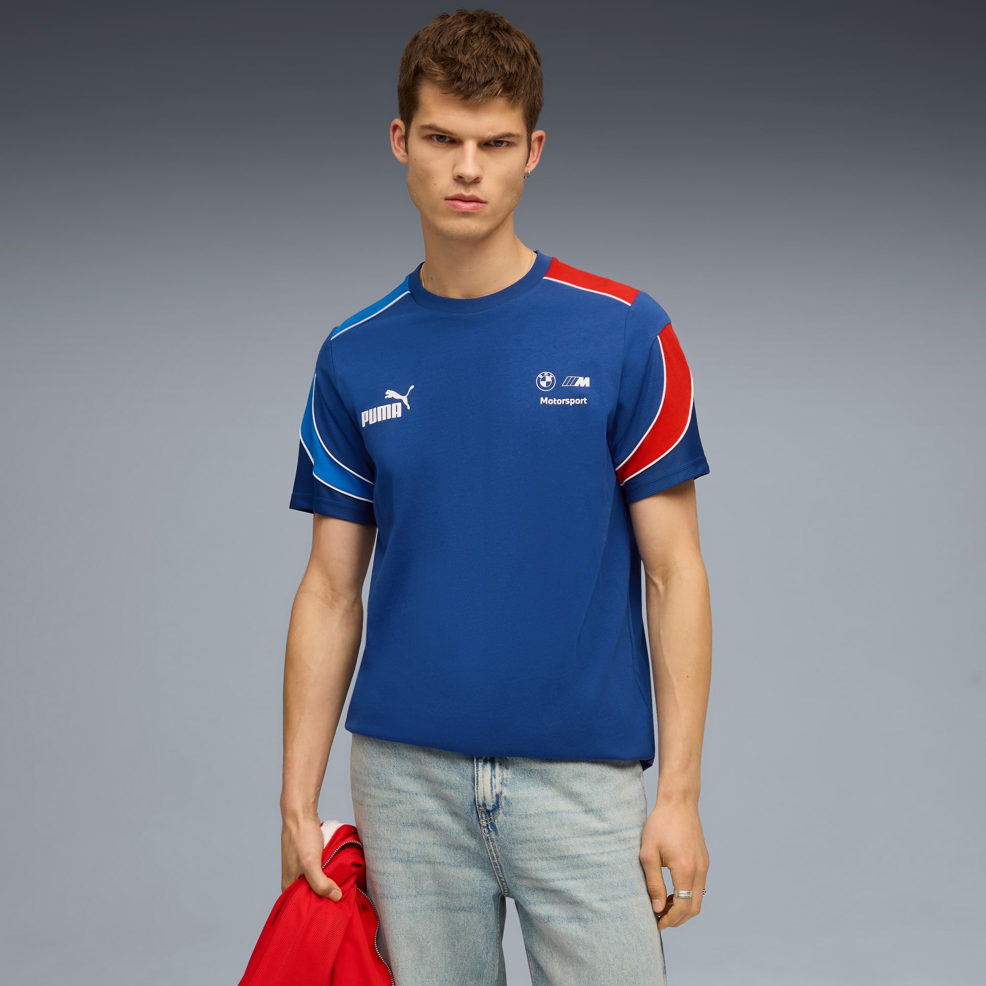 Un modèle pose sur fond gris, portant un t-shirt bleu, un jean clair et tenant une veste rouge.