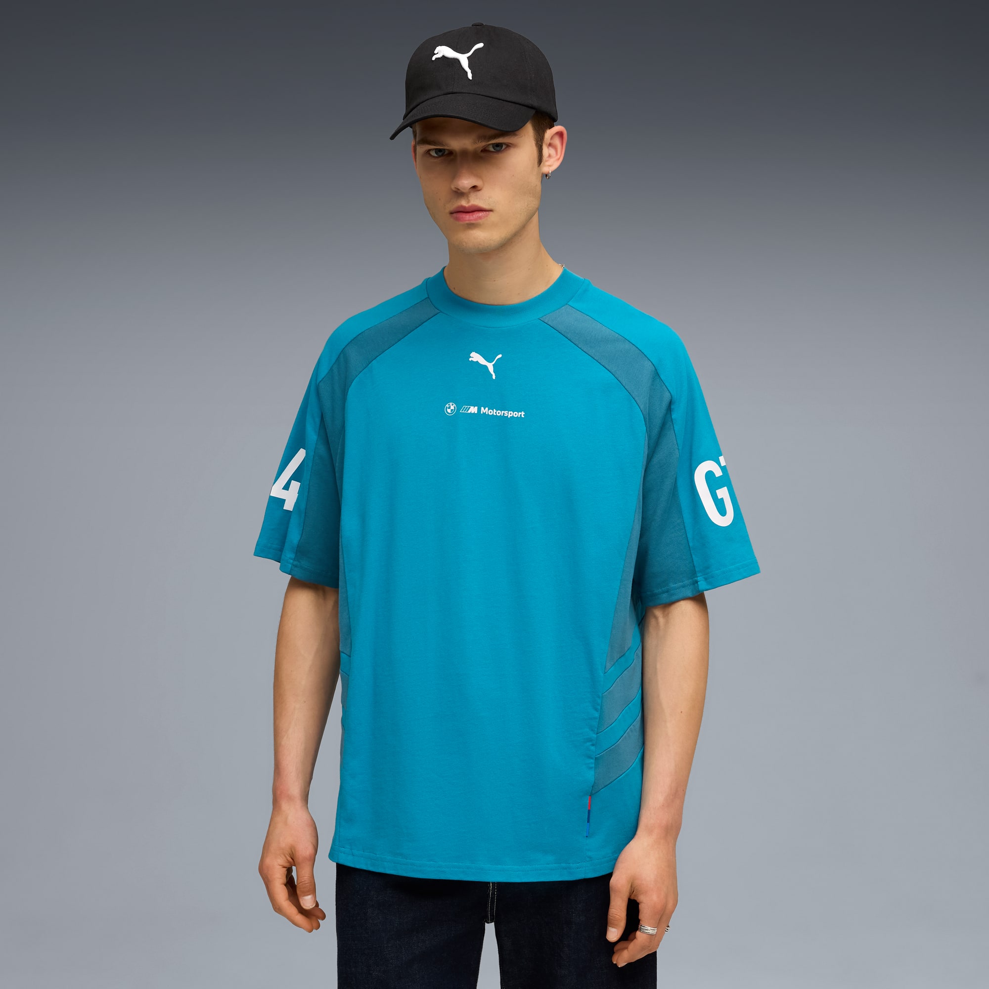 Un modello posa su sfondo grigio, indossa un cappellino, una t-shirt blu e jeans scuri.