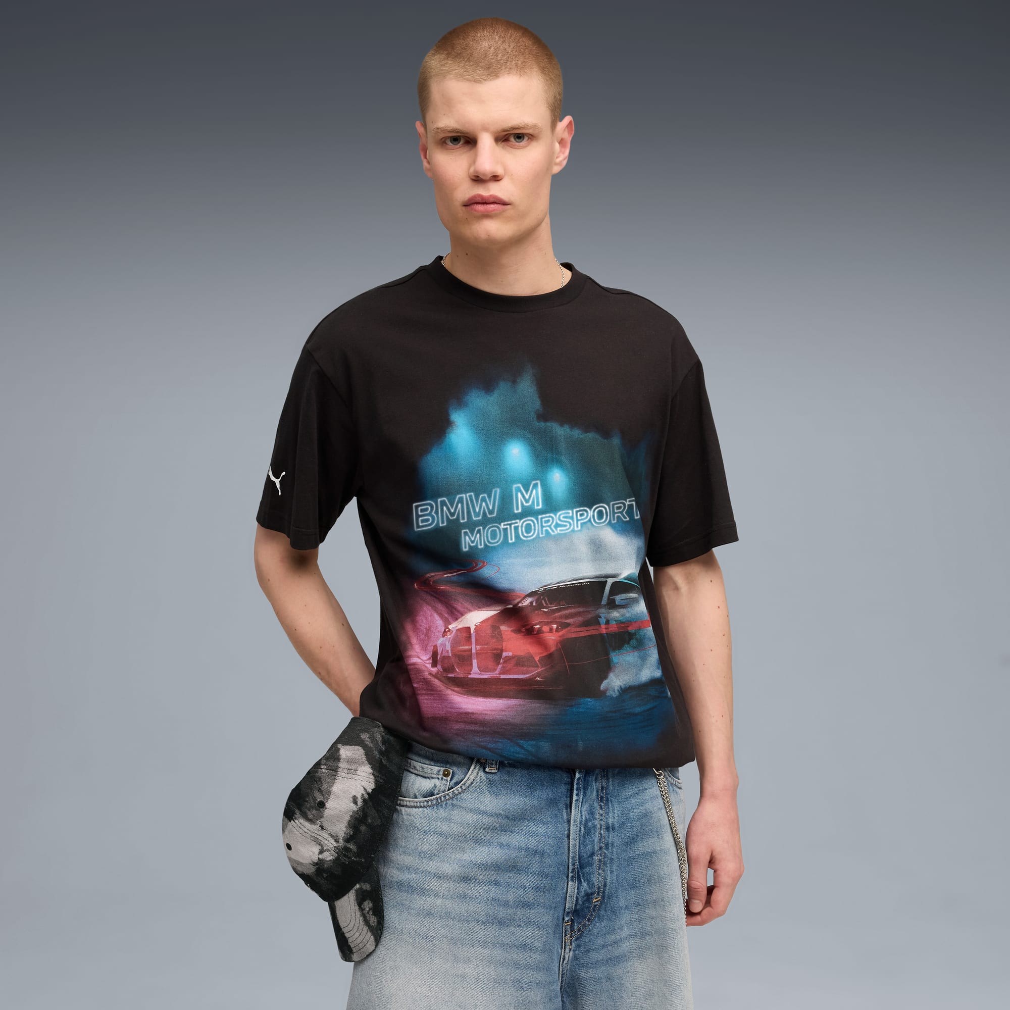 Een model poseert tegen grijs met een T-shirt met print, denim shorts en pet.