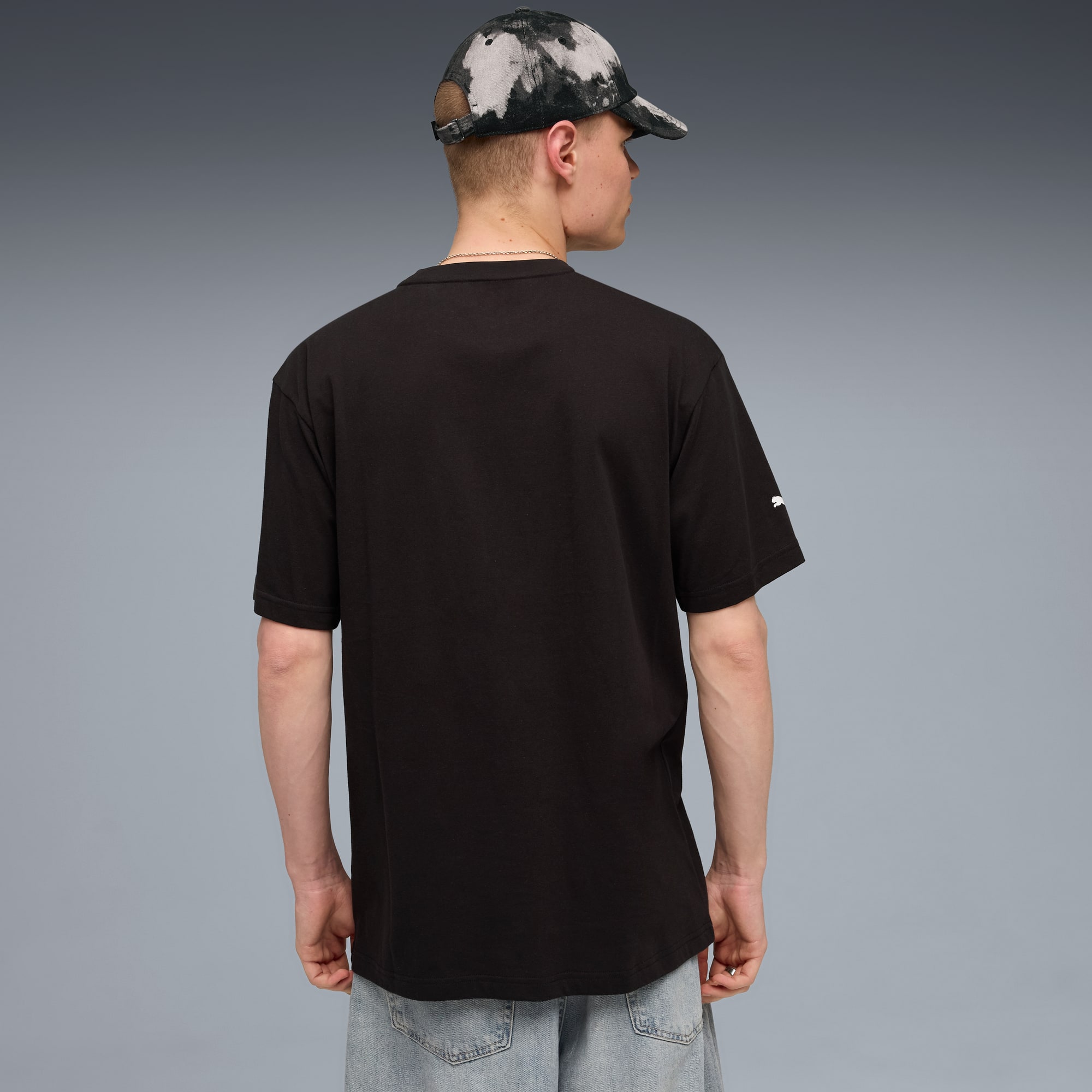 Een model staat met de rug naar de camera op een grijze achtergrond, met zwart t-shirt, lichte jeans en pet.