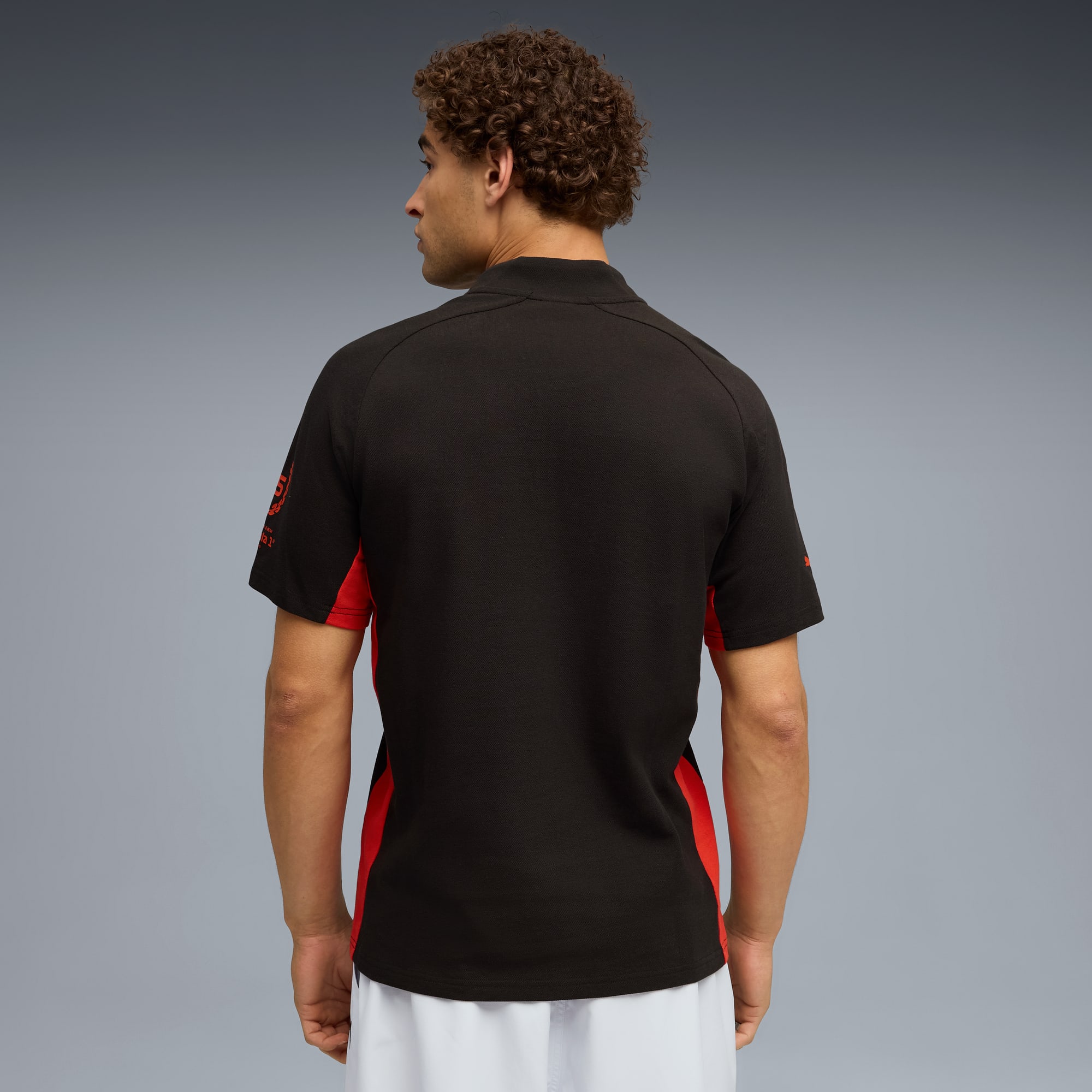Su sfondo grigio, un modello di spalle indossa una maglia nera e rossa con pantaloni bianchi.
