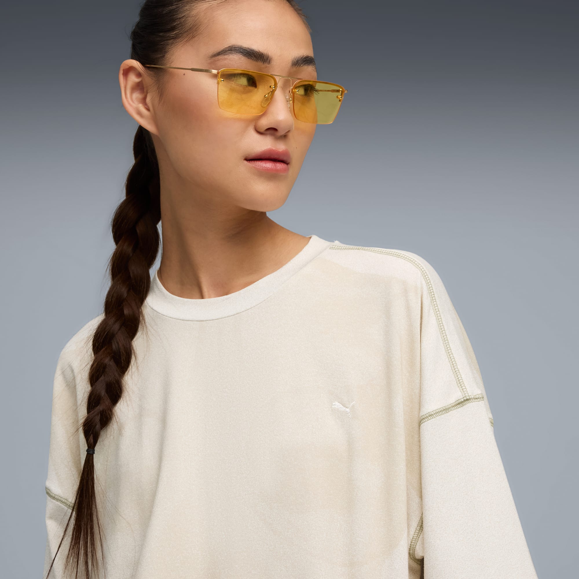 Model met vlecht poseert tegen grijs, draagt top en geelgetinte bril.