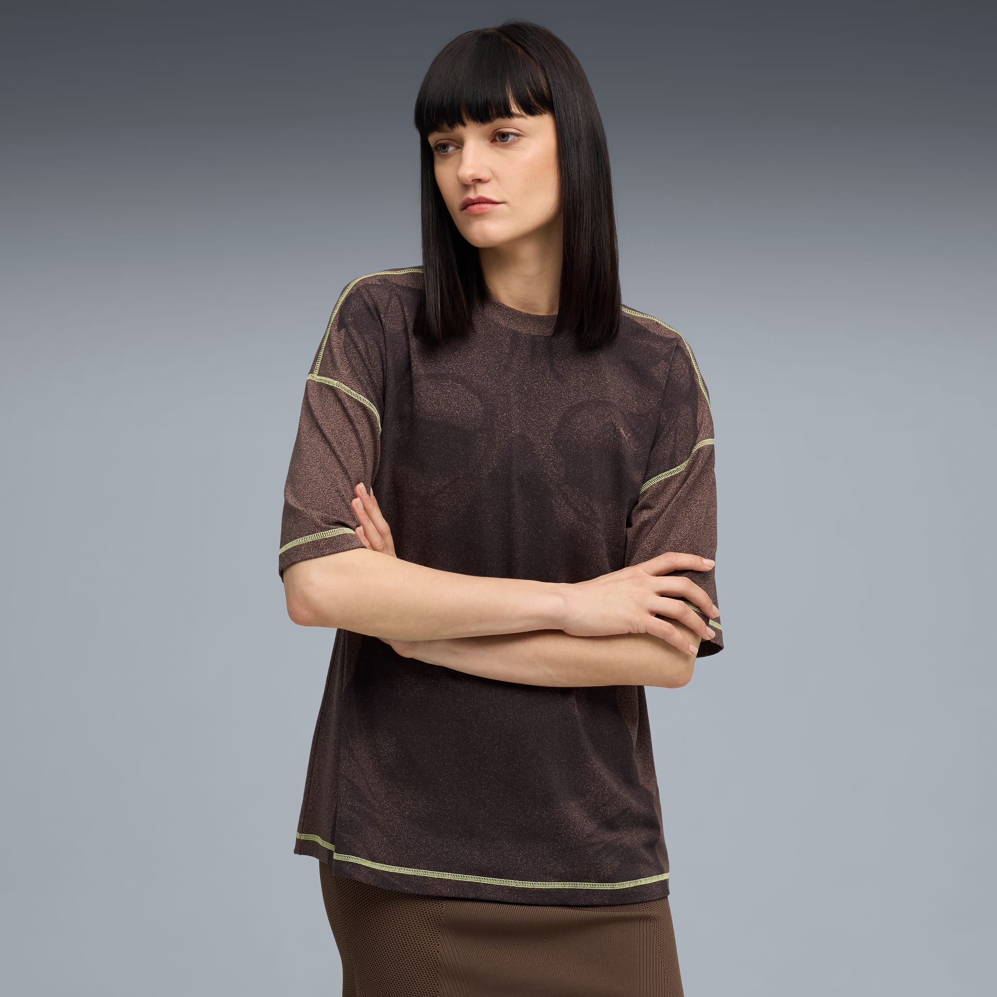 Vor grauem Hintergrund steht ein Model mit verschränkten Armen, braunem Shirt und Rock.