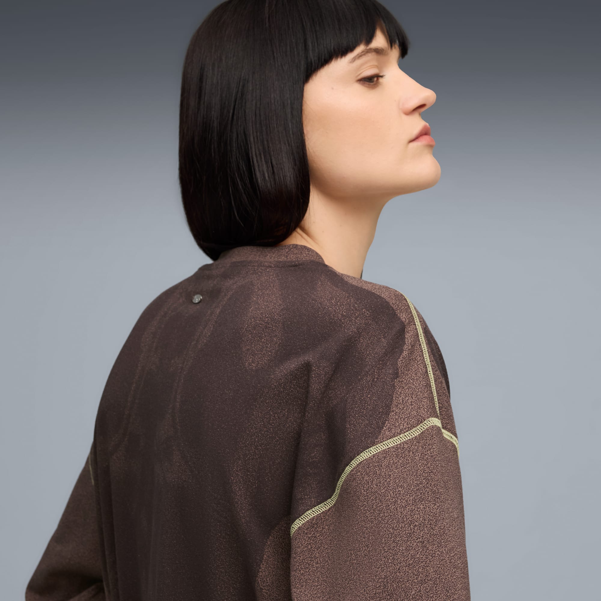 Ein Model in einem braunen Oberteil mit gelben Nähten posiert vor grauem Hintergrund.
