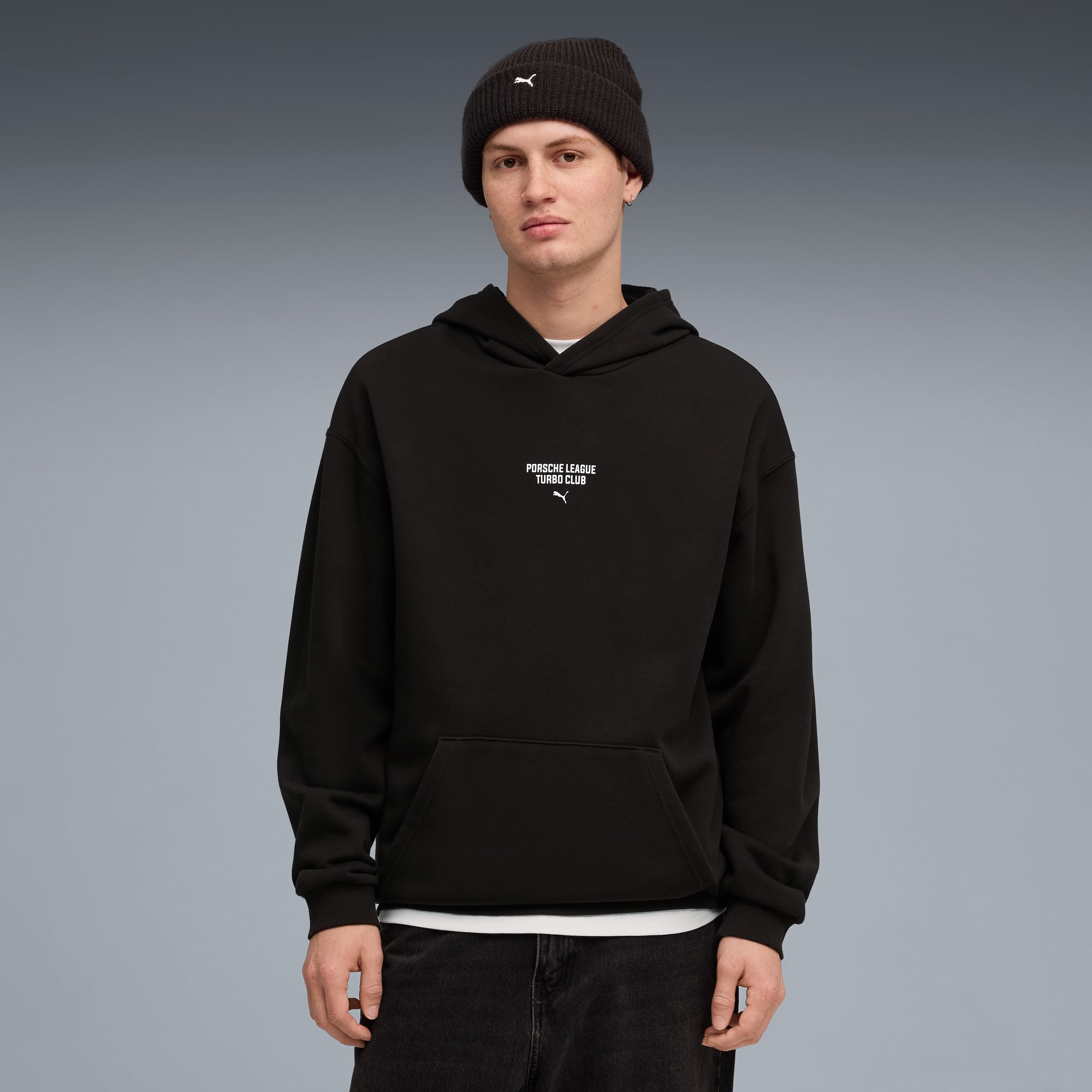 Ein Model steht vor grauem Hintergrund mit Mütze, schwarzem Hoodie und Jeans.