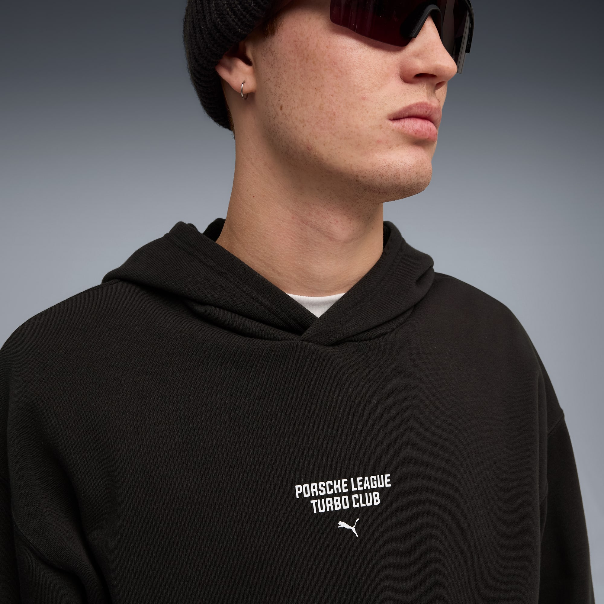 Ein Model trägt einen schwarzen Hoodie mit Porsche- und Puma-Logos vor grauem Hintergrund.