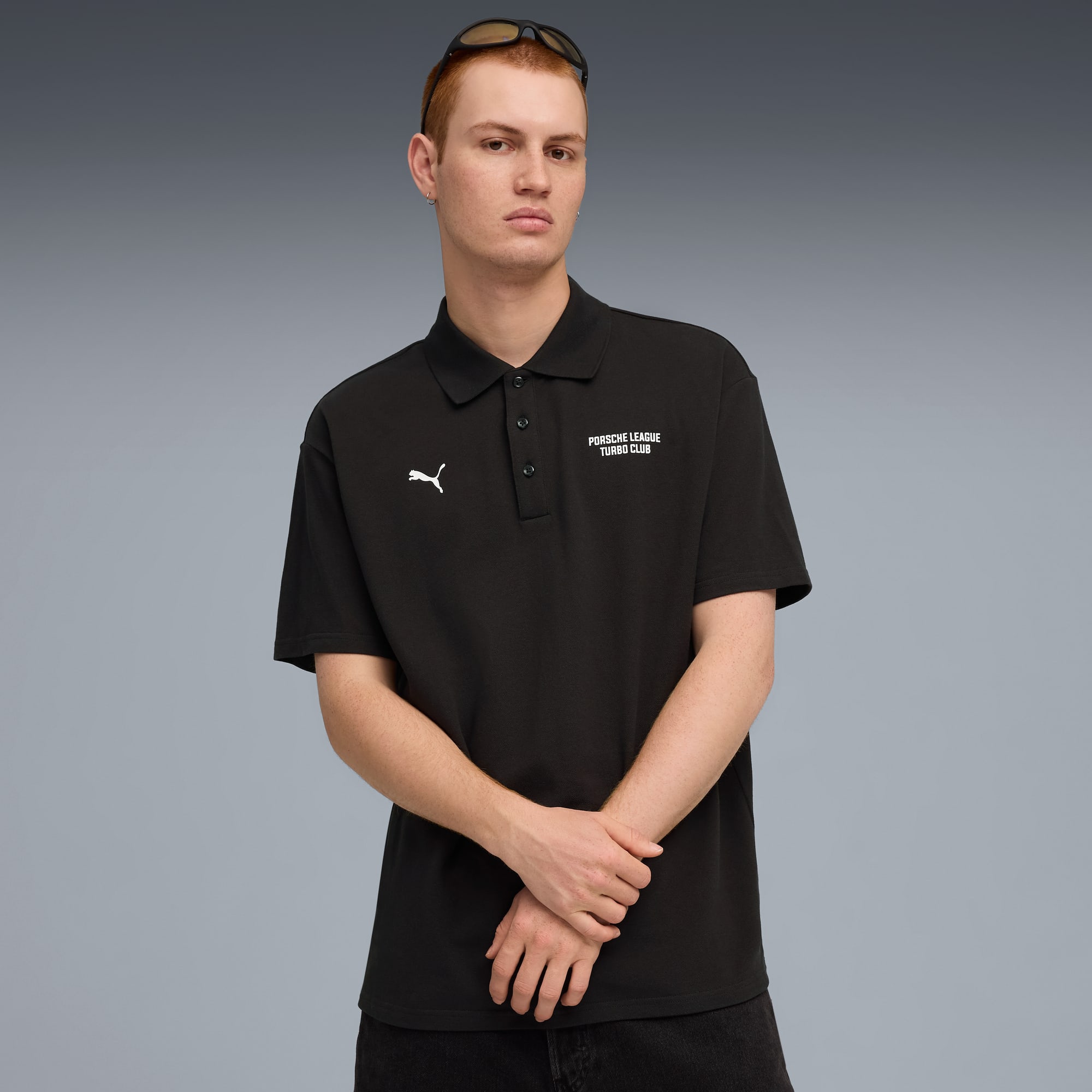 Vor grauem Hintergrund posiert ein Model in schwarzem Poloshirt mit Logos und dunkler Hose.