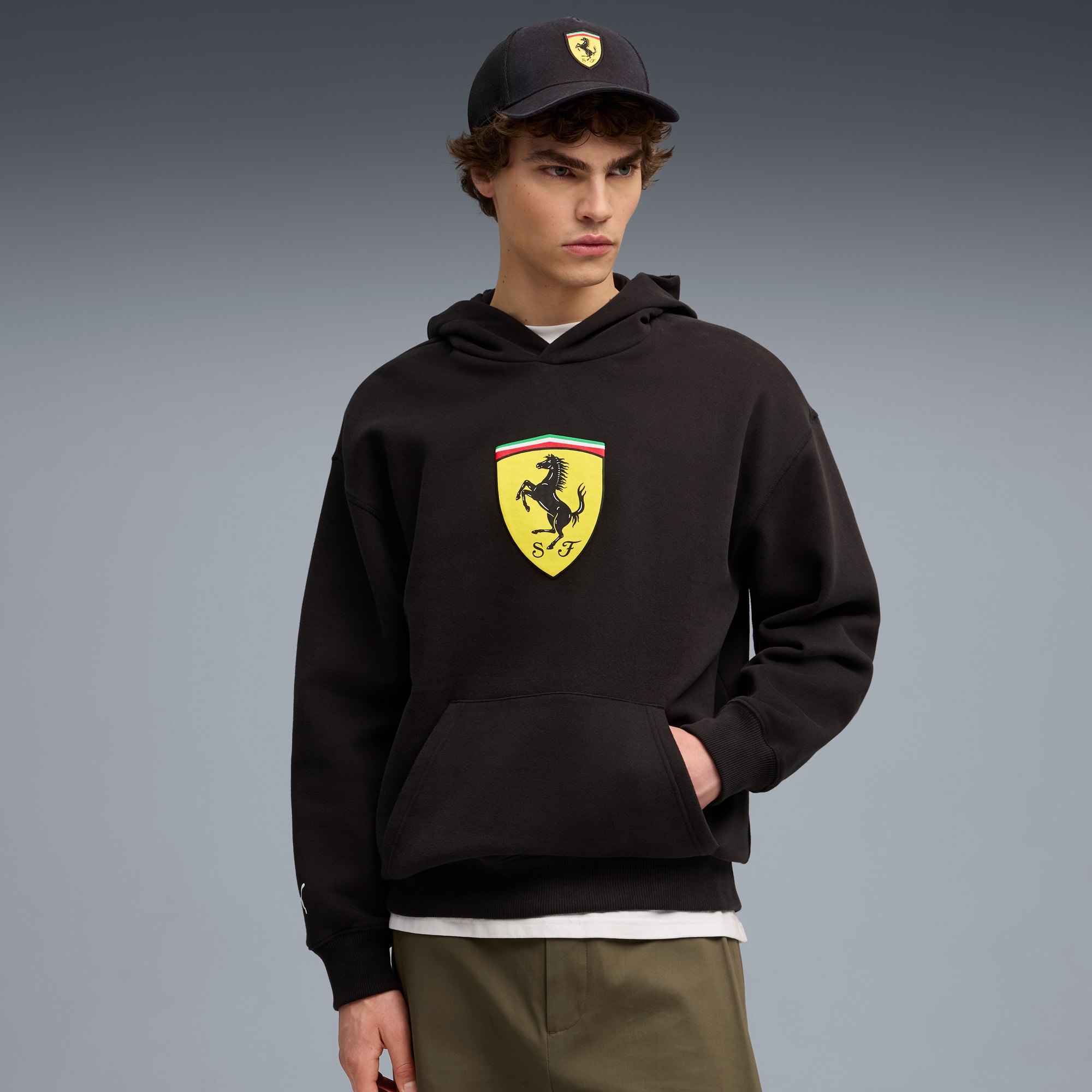 Ein Model posiert vor grauem Hintergrund mit Kappe und schwarzem Hoodie mit Abzeichen.