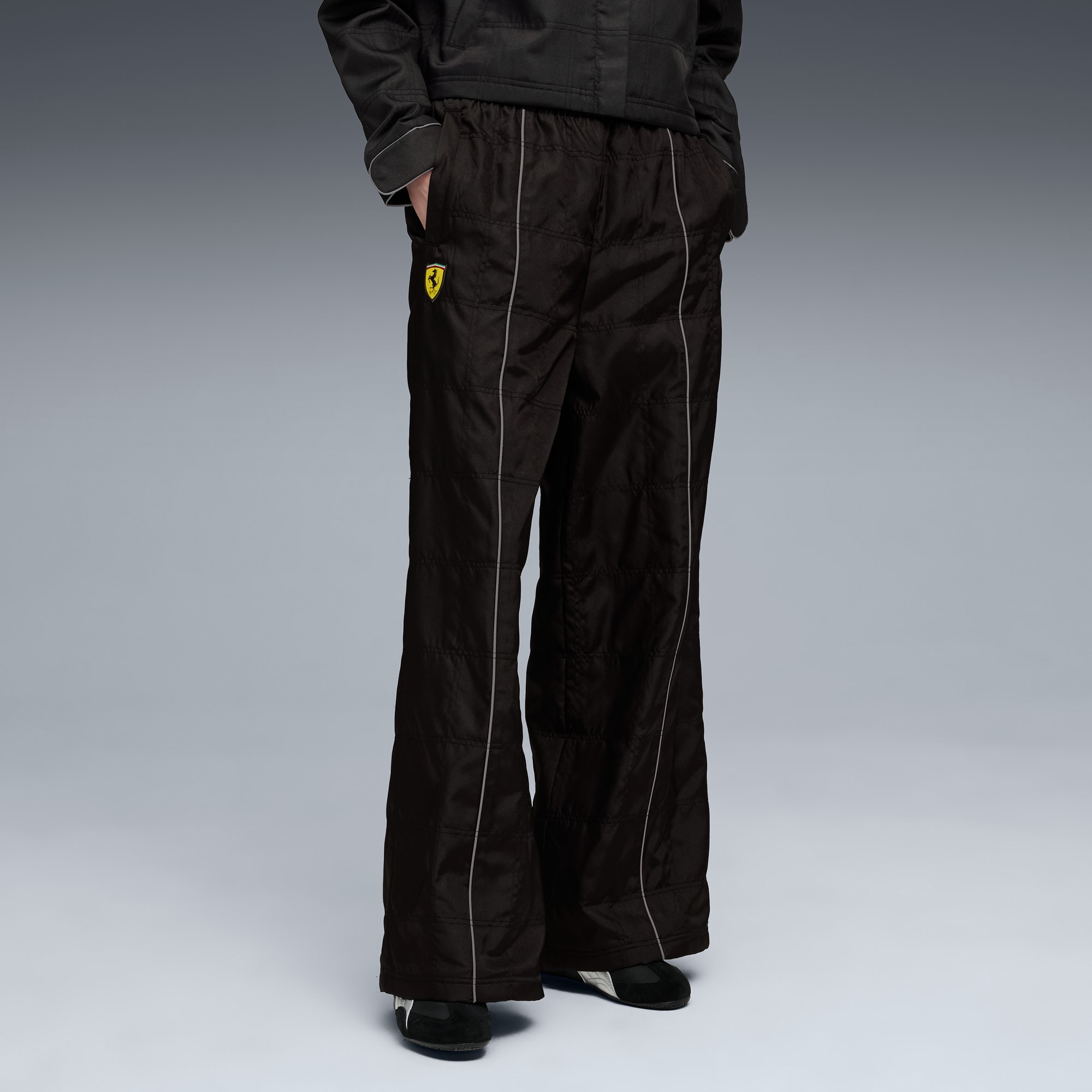 Vor grauem Hintergrund trägt ein Model eine schwarze Hose mit hellem Besatz und Logo.