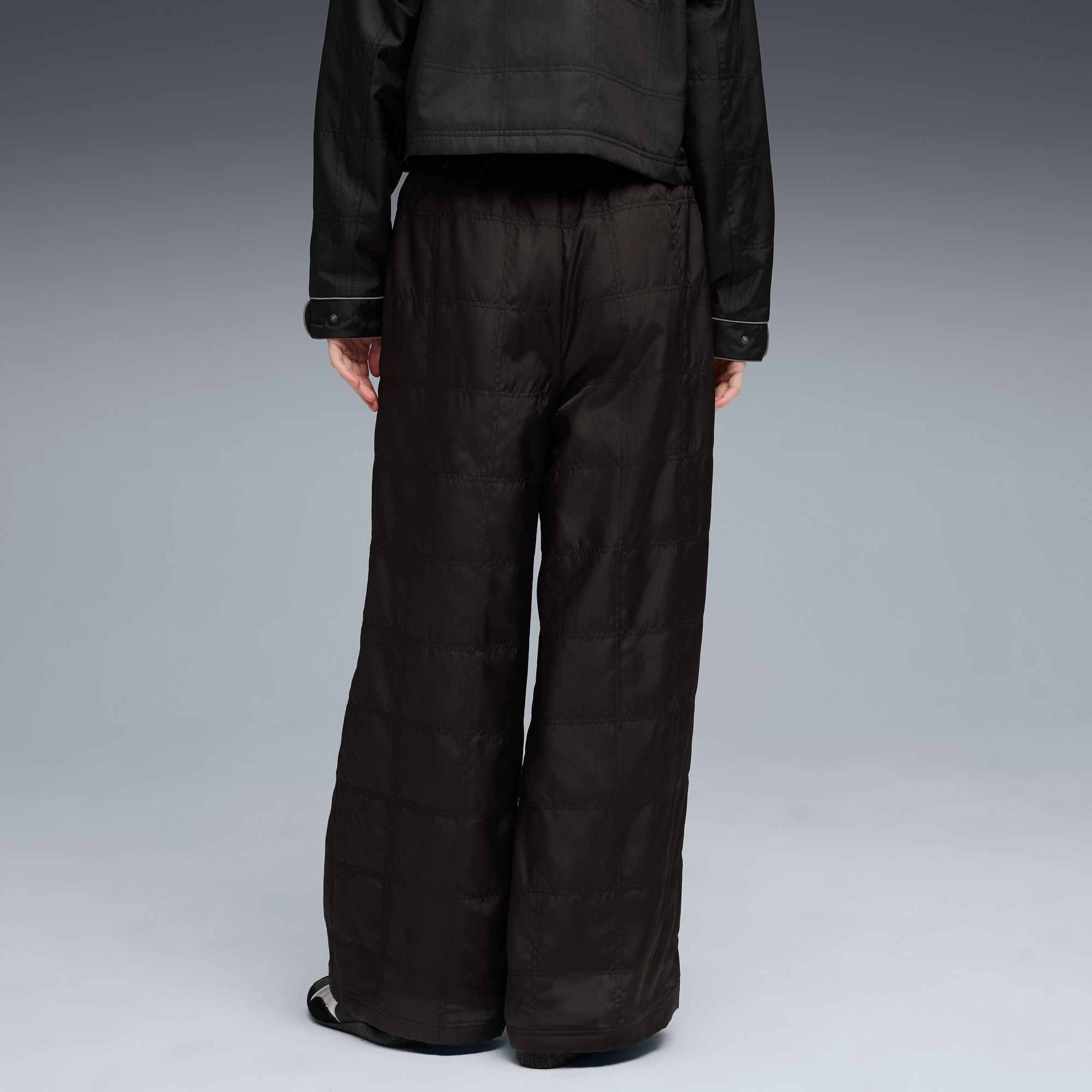 Ein Model vor grauem Hintergrund trägt eine schwarze Jacke und gesteppte, weit geschnittene Hosen.