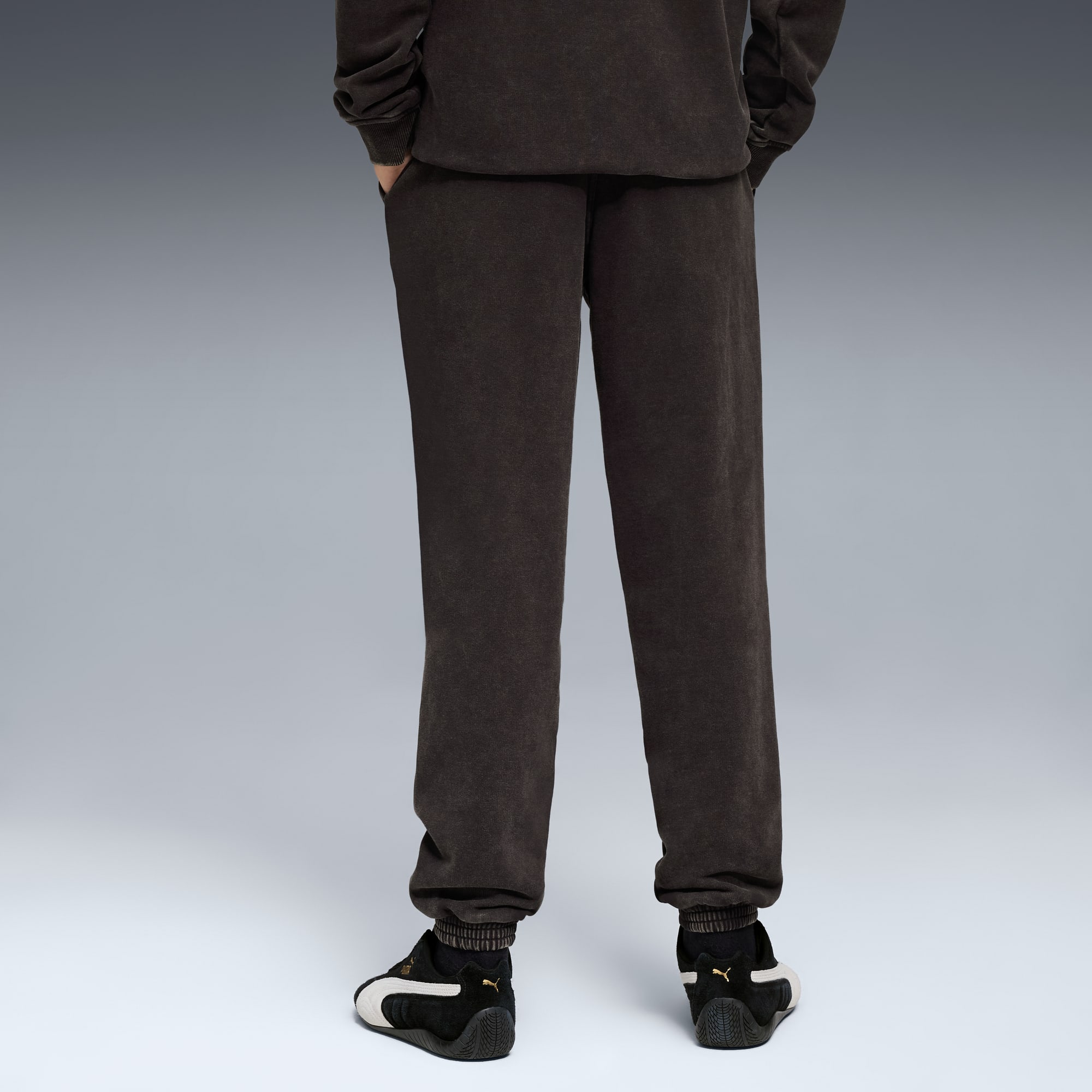 Ein Model mit Händen in den Taschen trägt braune Jogginghose und schwarz-weiße Turnschuhe auf Grau.